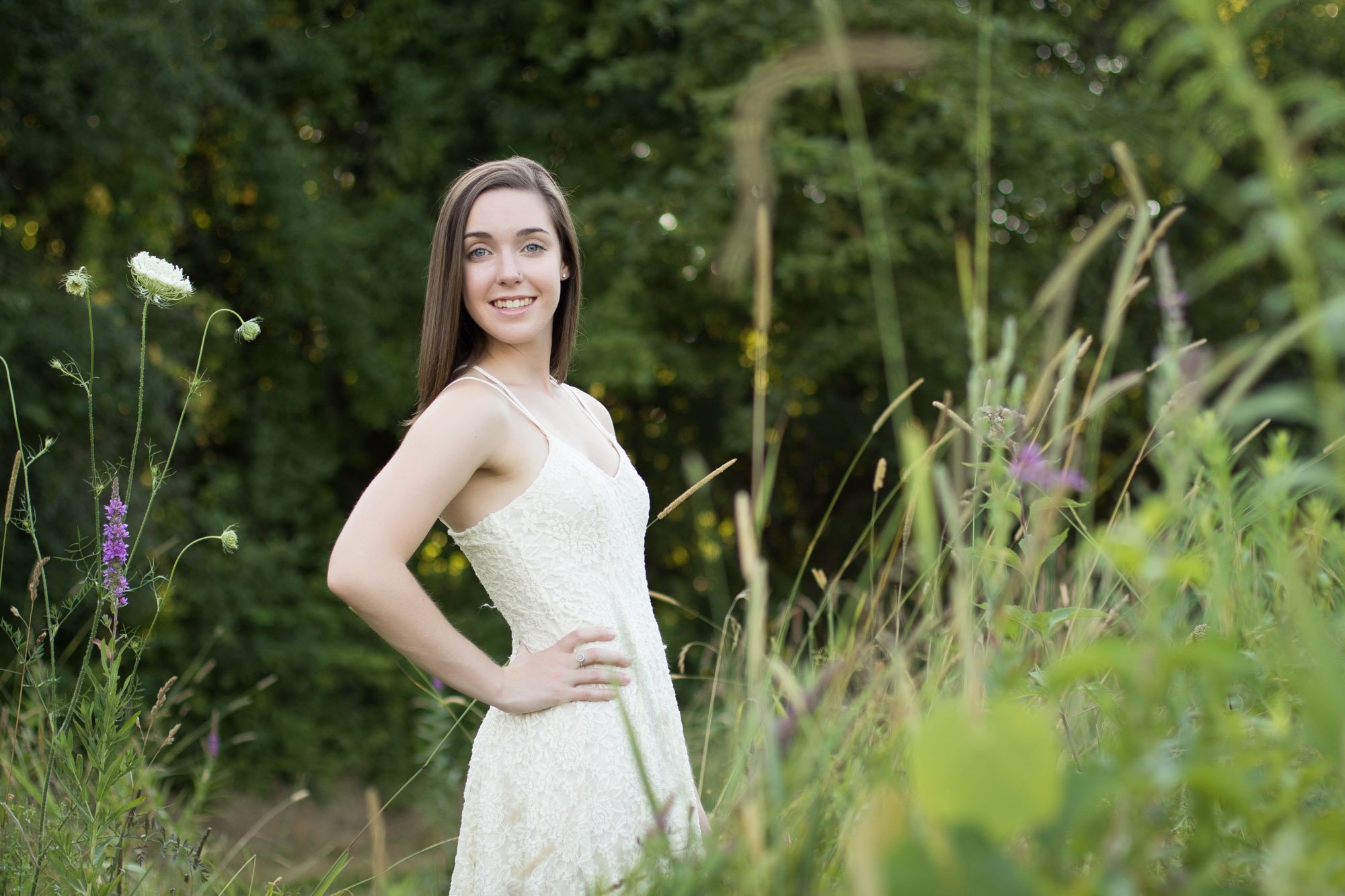 Young woman in a white lace dress standing in a green, wooded area with wildflowers.