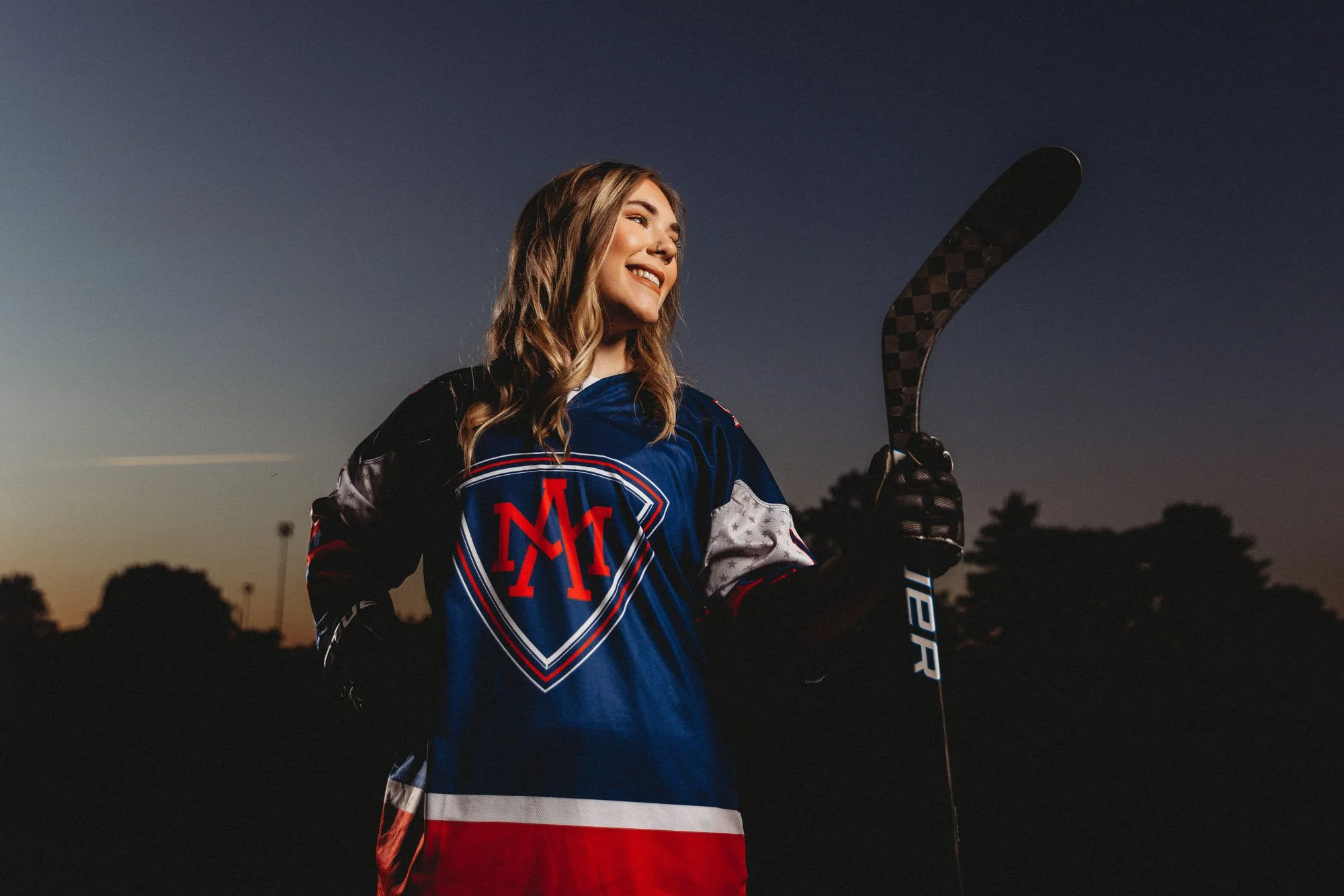 A woman in a New York Rangers hockey jersey holding a hockey stick, standing outdoors during sunset.