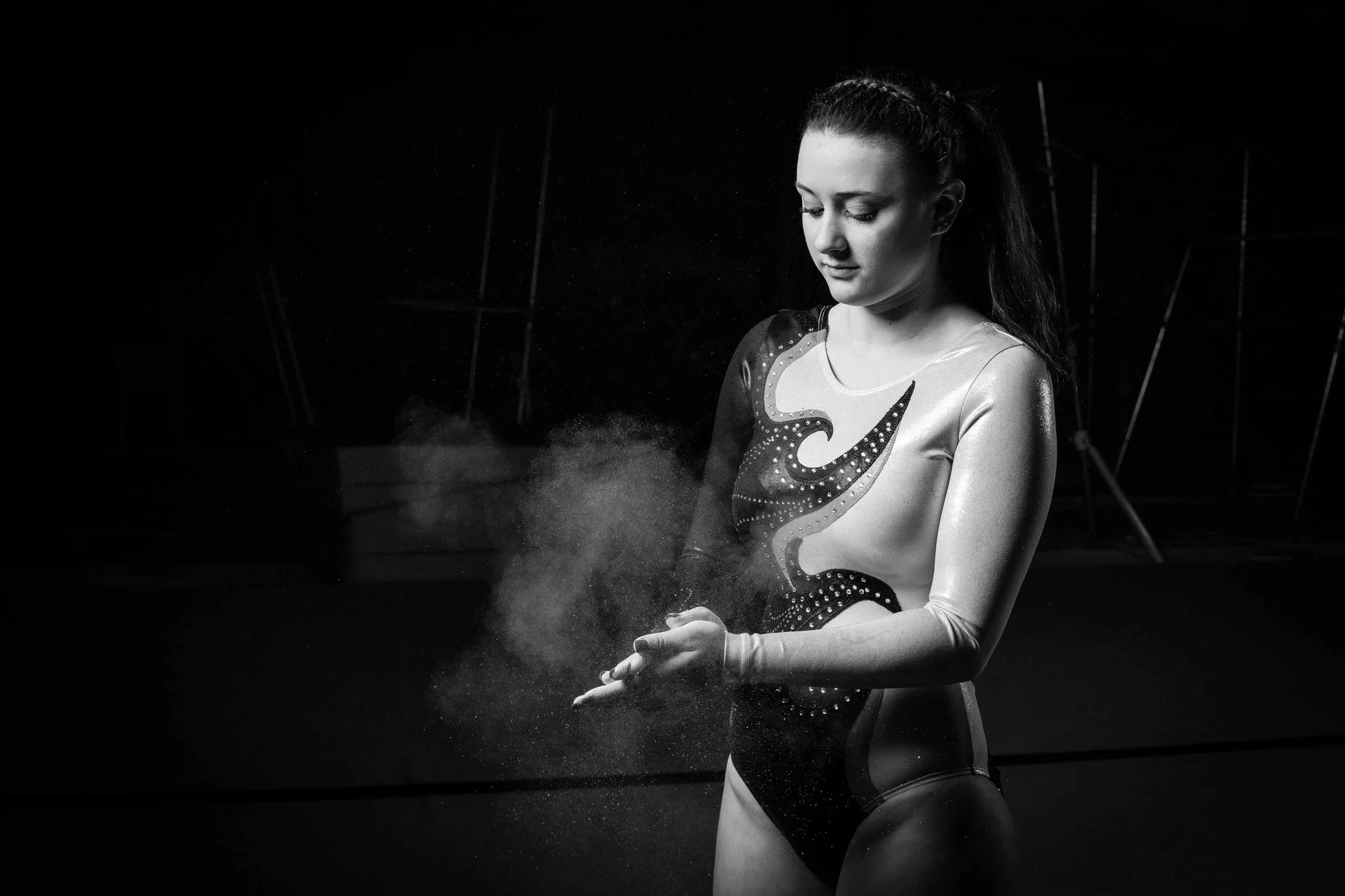 A female gymnast with a focused expression, dressed in a sparkly leotard, holds chalk dust in her hands against a dark background.