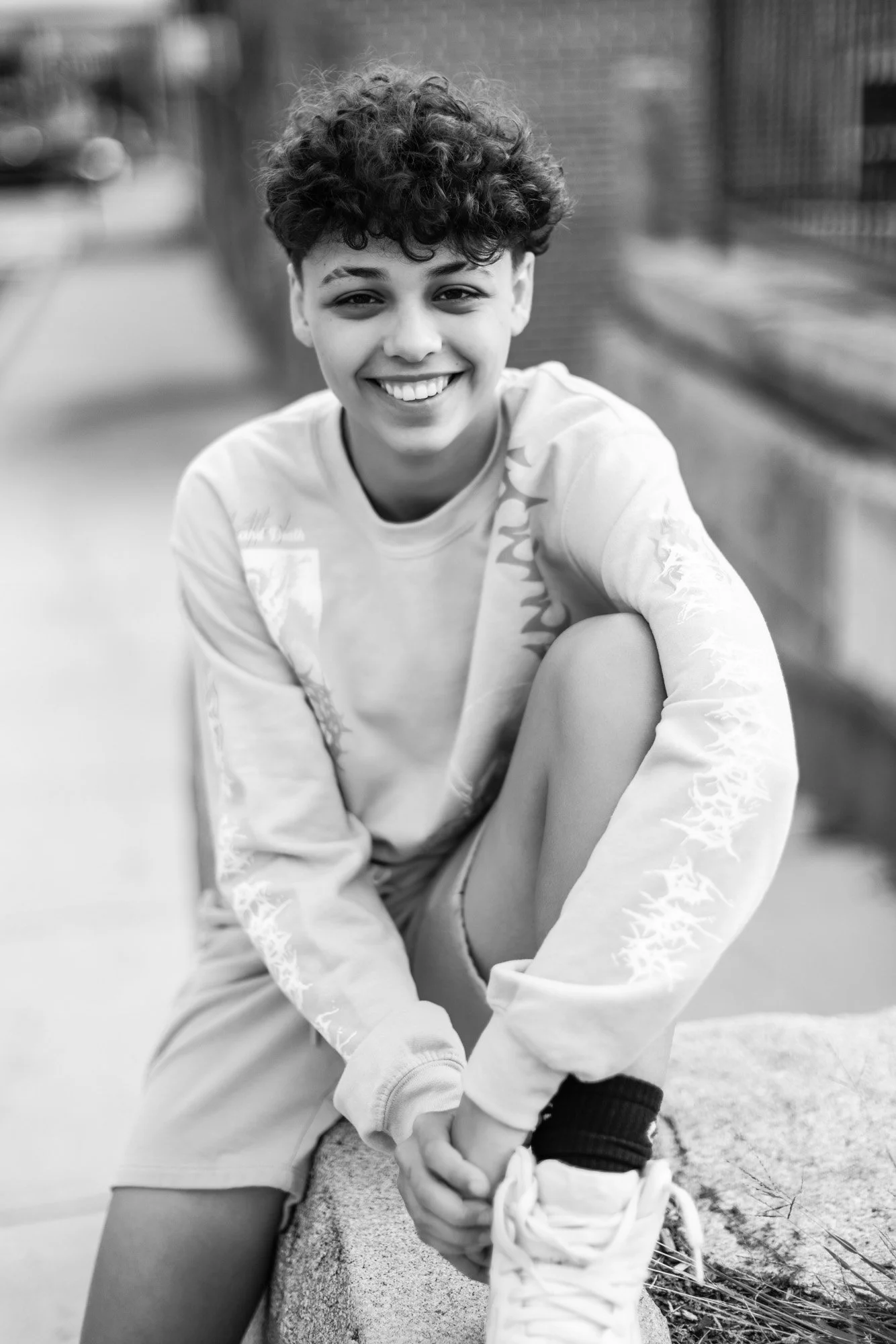 A smiling young person with curly hair sitting on a concrete ledge outdoors, tying their shoelace, in black and white.