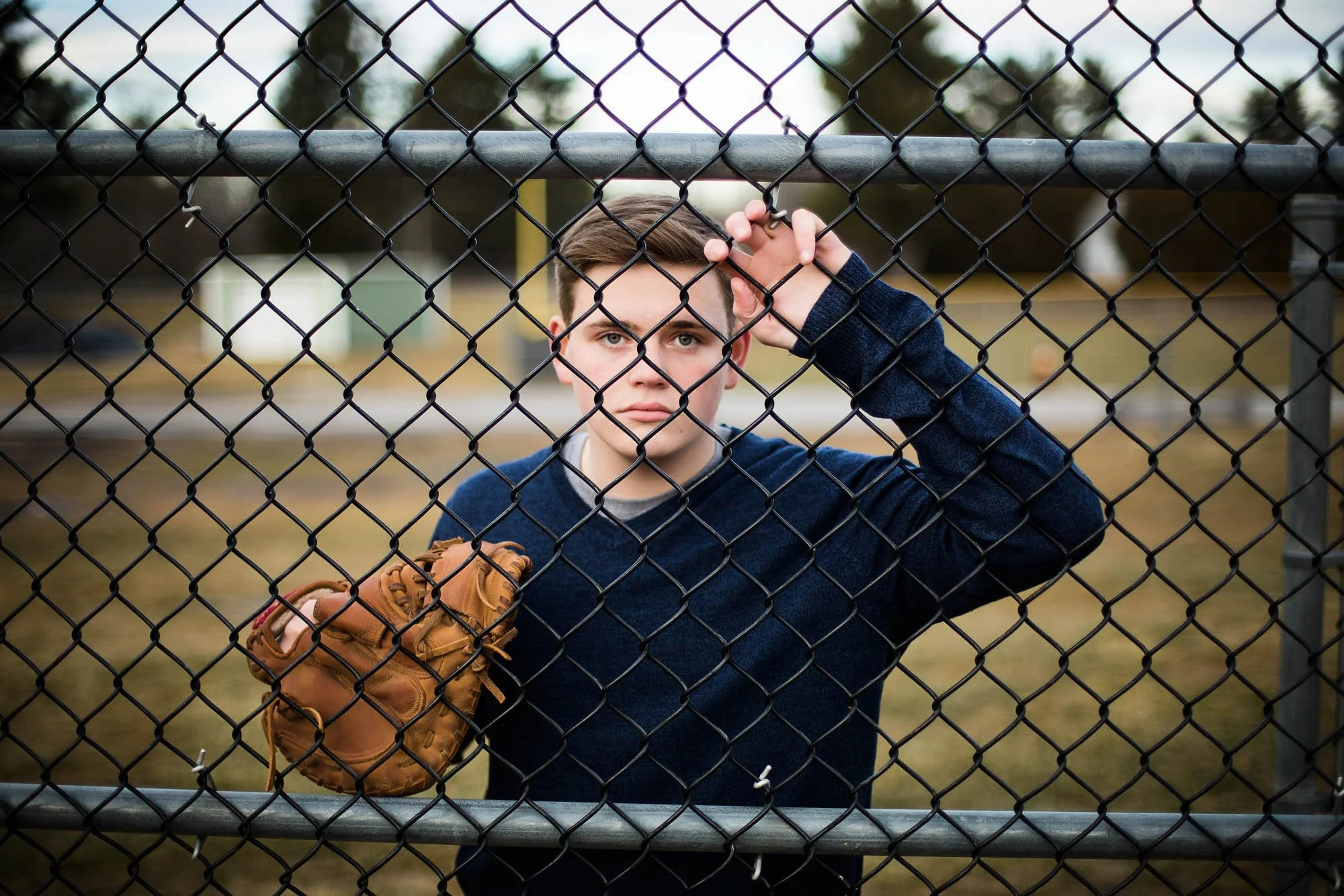 A young man with short brown hair and wearing a dark blue hoodie stands behind a chain-link fence at a baseball field, holding a tan baseball glove in his left hand.