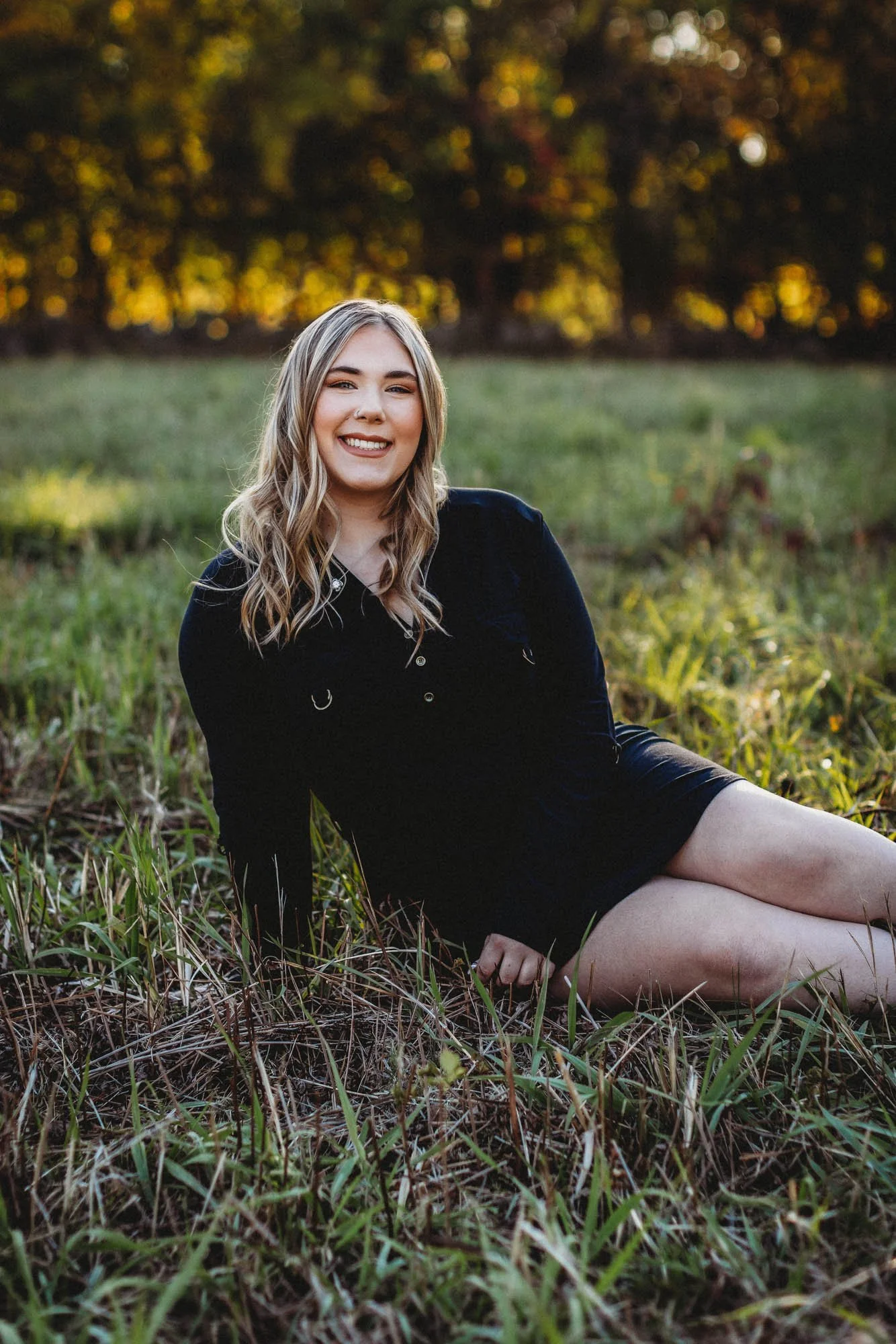 A young woman with blonde hair, wearing a black dress, sitting on the grass in a field during sunset, smiling at the camera.