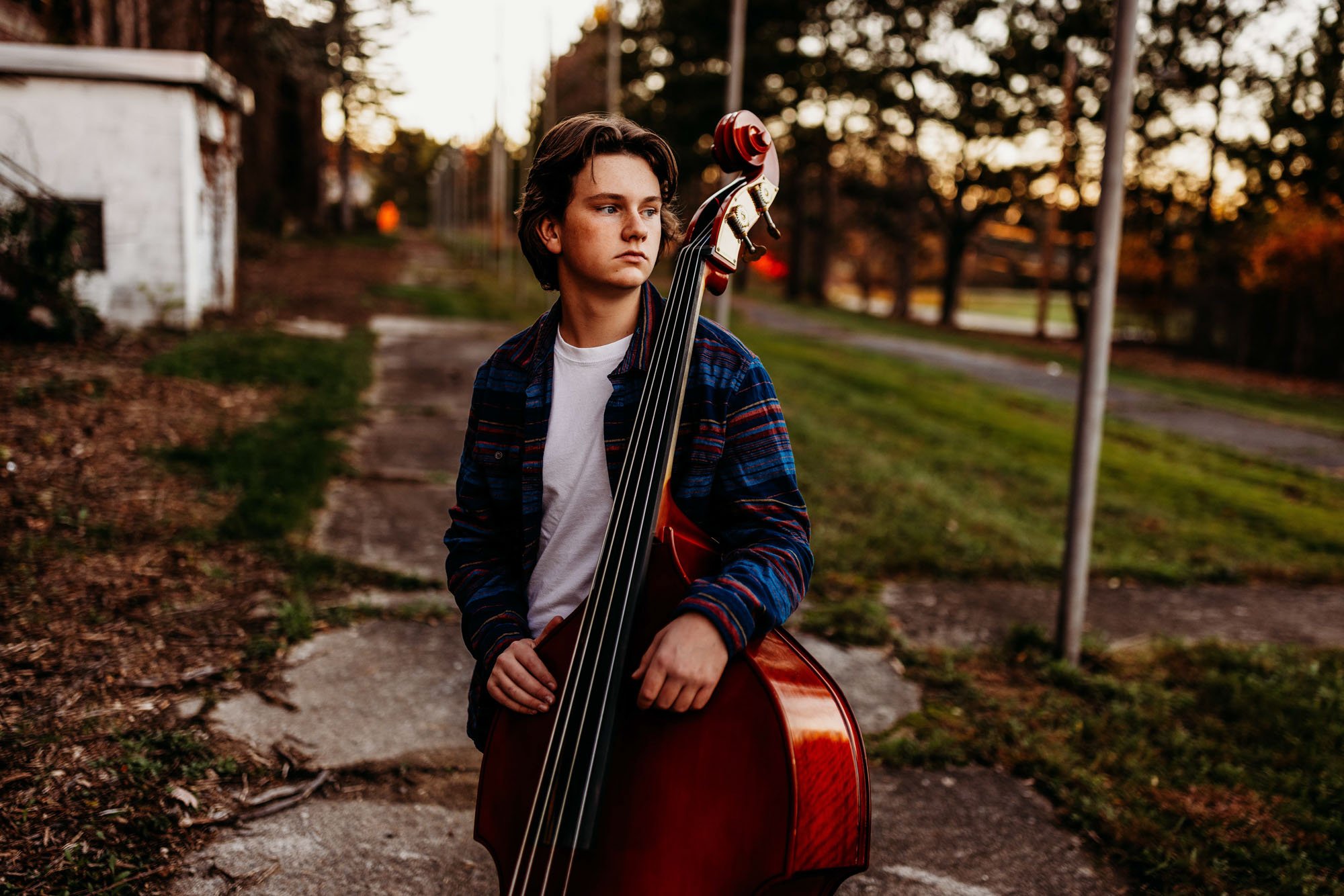 A young person with brown hair stands outdoors holding a double bass, looking to the side. The background features a sidewalk, grass, and trees during sunset.