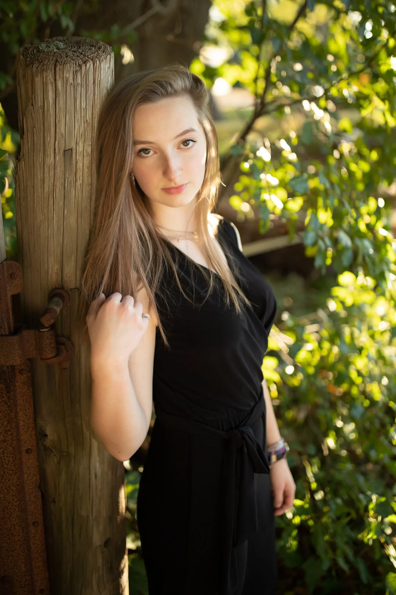 A young woman with long, light brown hair, wearing a black sleeveless top and black pants, standing outdoors by a wooden post among greenery, looking at the camera.
