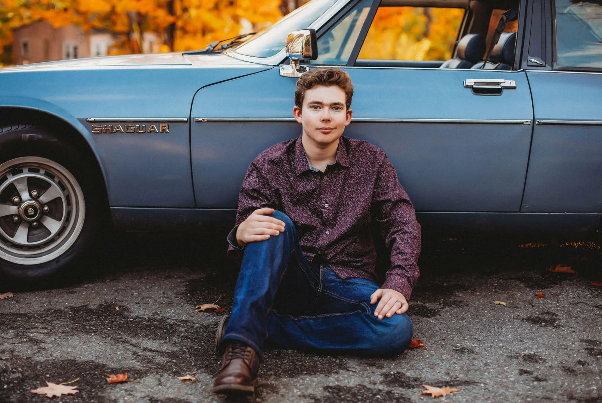 A young man sitting on the ground in front of a blue vintage Jaguar car with autumn leaves on the ground and trees in the background.
