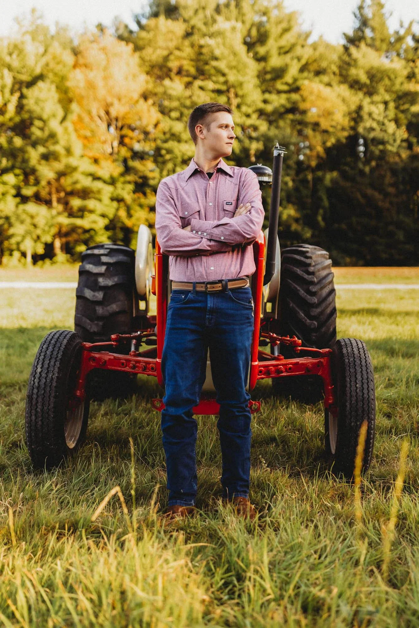 A young man stands with arms crossed in front of a red tractor in a grassy field, with trees in the background during autumn.