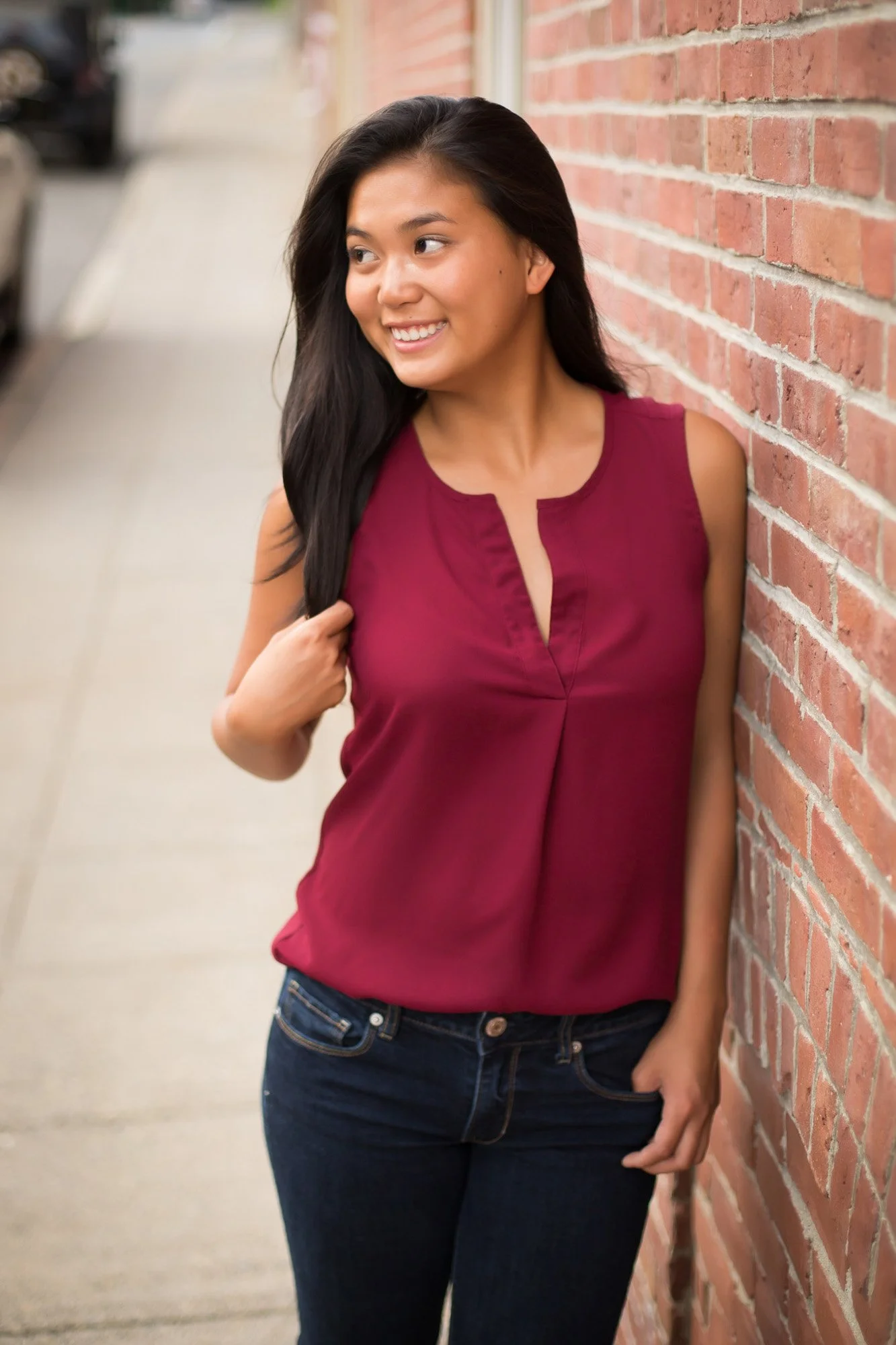 A young woman with long black hair smiling and leaning against a brick wall on a sidewalk in an urban setting.