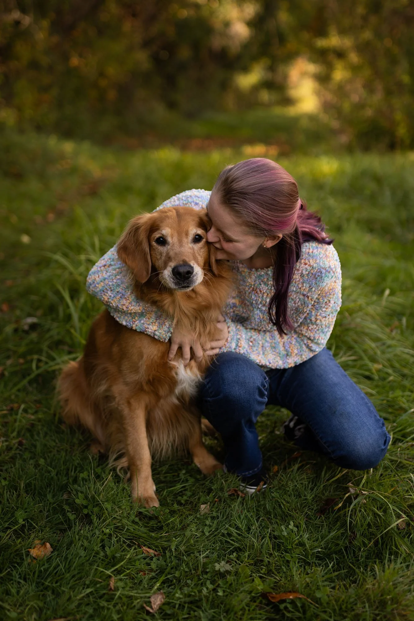 A woman hugging a golden retriever dog outdoors in a grassy area surrounded by trees.