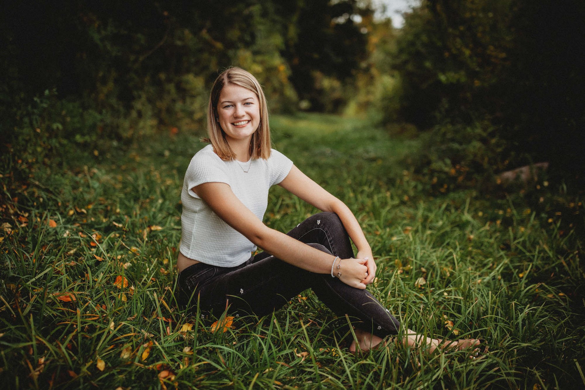A young woman with shoulder-length blonde hair, wearing a white short-sleeve top and black jeans, sitting on lush green grass in a wooded area during daytime, smiling at the camera.