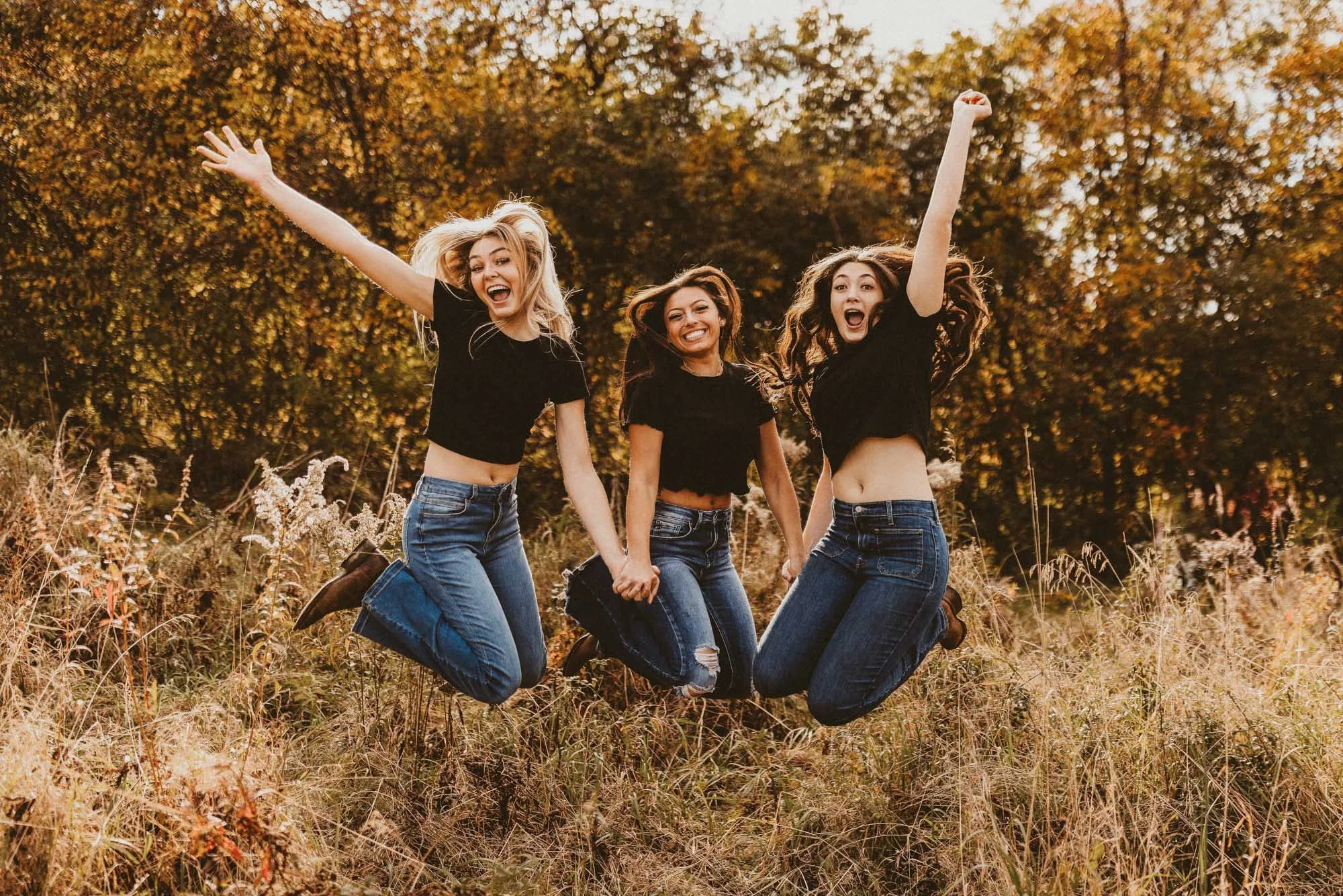 Three young women jumping in a grassy field during autumn, with trees in the background, wearing black t-shirts and jeans, smiling and holding hands.