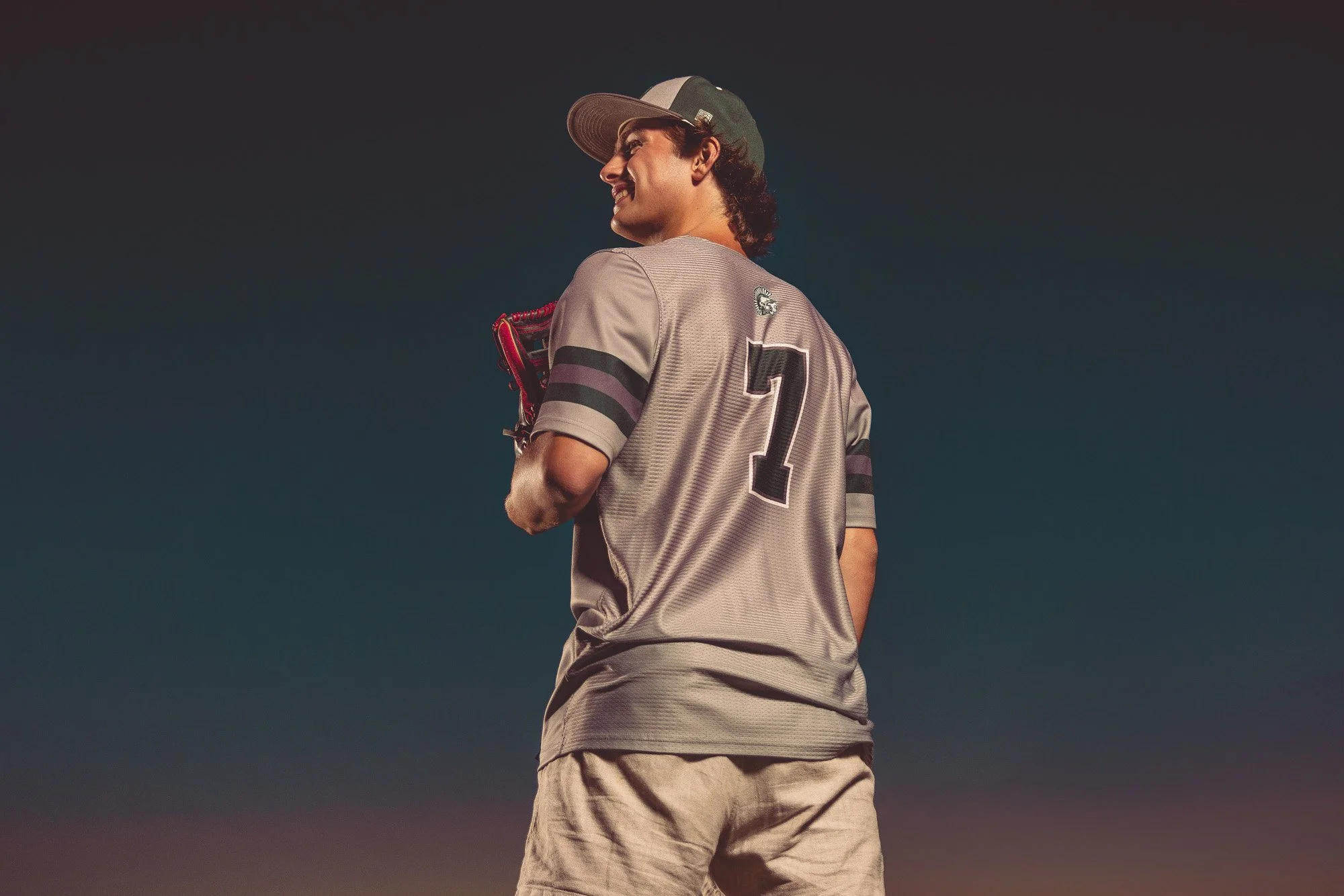 A young man in a baseball uniform with the number seven on the back, holding a baseball glove, standing outdoors during sunset or twilight.