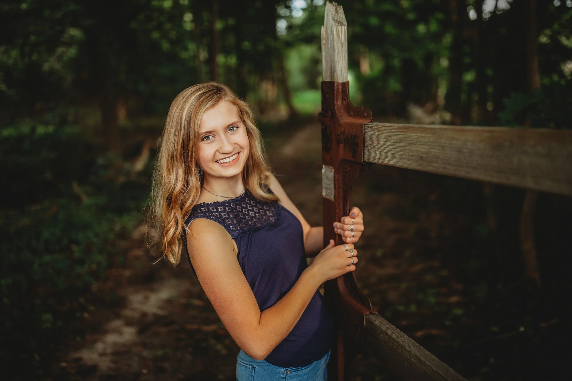 A young woman with blonde hair and a navy blue sleeveless top standing near a rustic wooden fence in a wooded area.