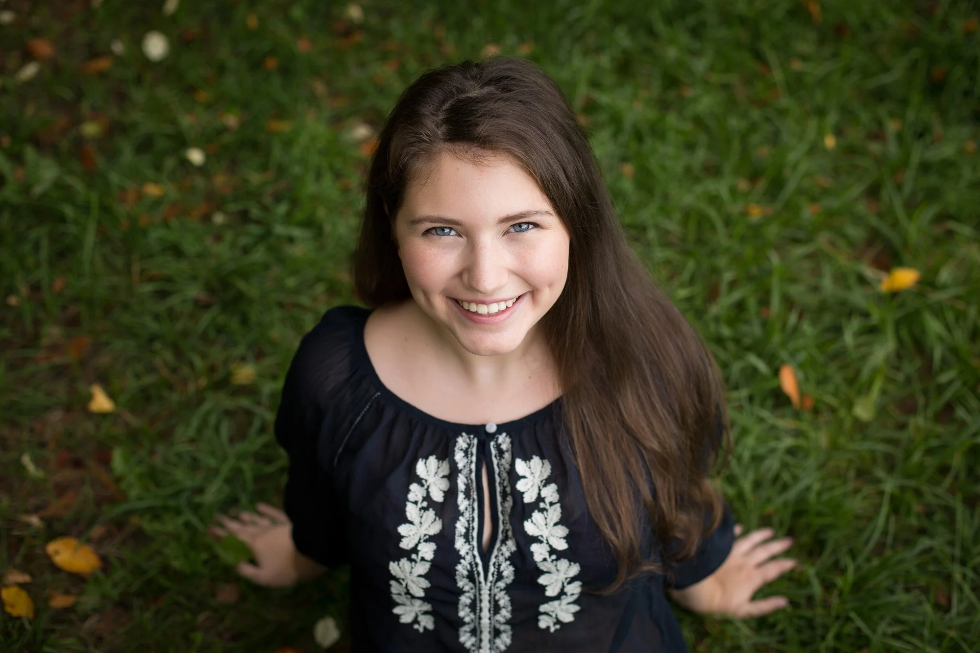 Young woman with long brown hair and blue eyes smiling, sitting on grass surrounded by fallen leaves, wearing a black top with white floral embroidery.