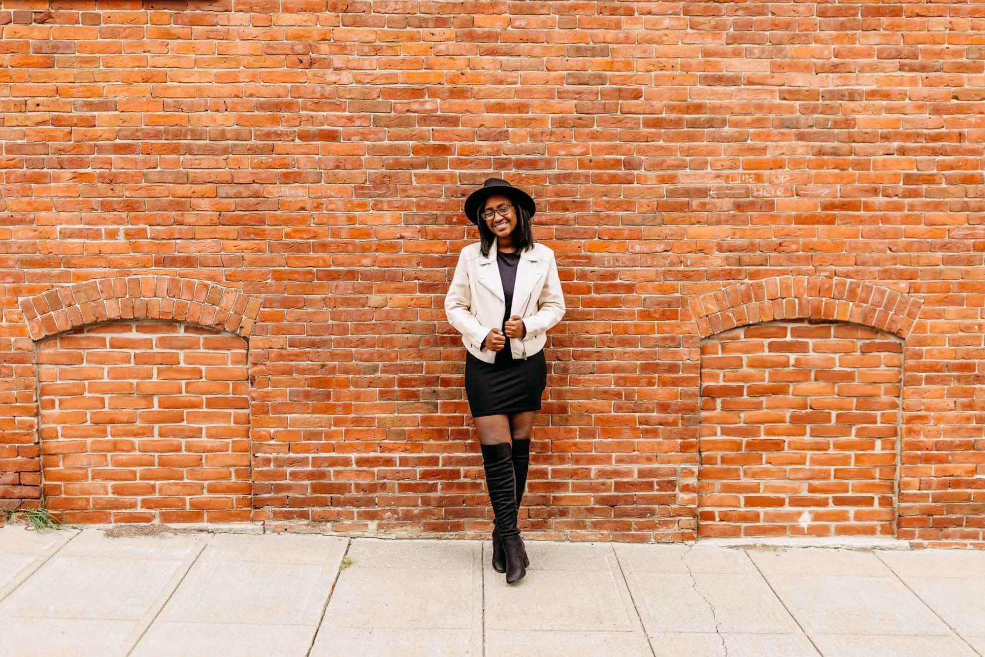 A woman dressed in black with a white jacket and black hat, smiling, standing against a brick wall.