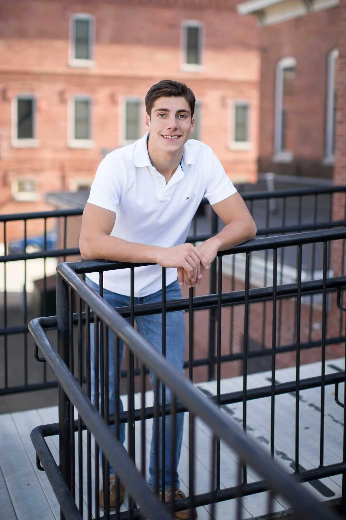 A young man with dark hair, wearing a white polo shirt and blue jeans, smiling and leaning on a black metal railing on a balcony with red brick buildings in the background.