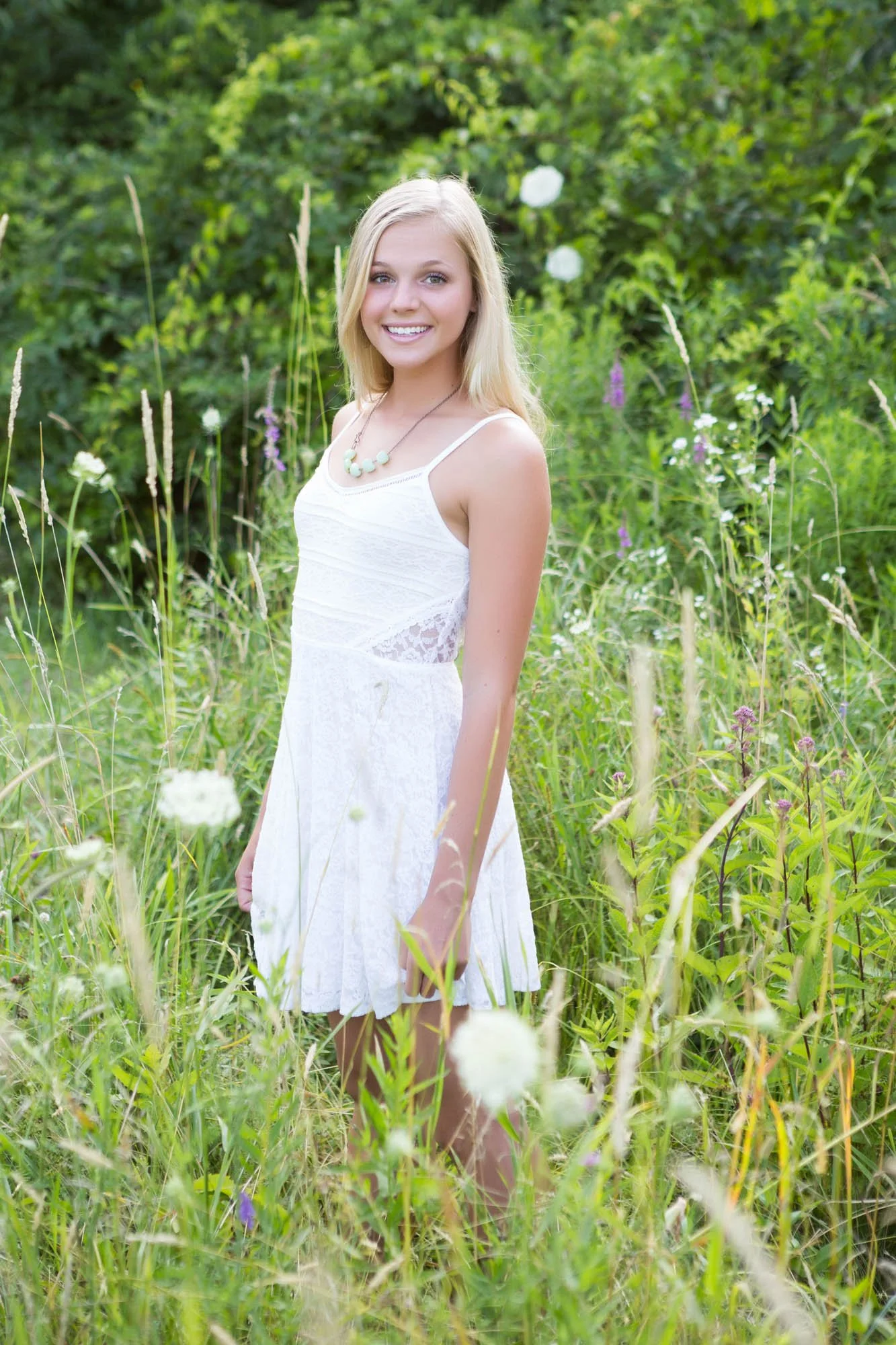 A young woman with blonde hair in a white dress standing in a lush green field with wildflowers, smiling at the camera.