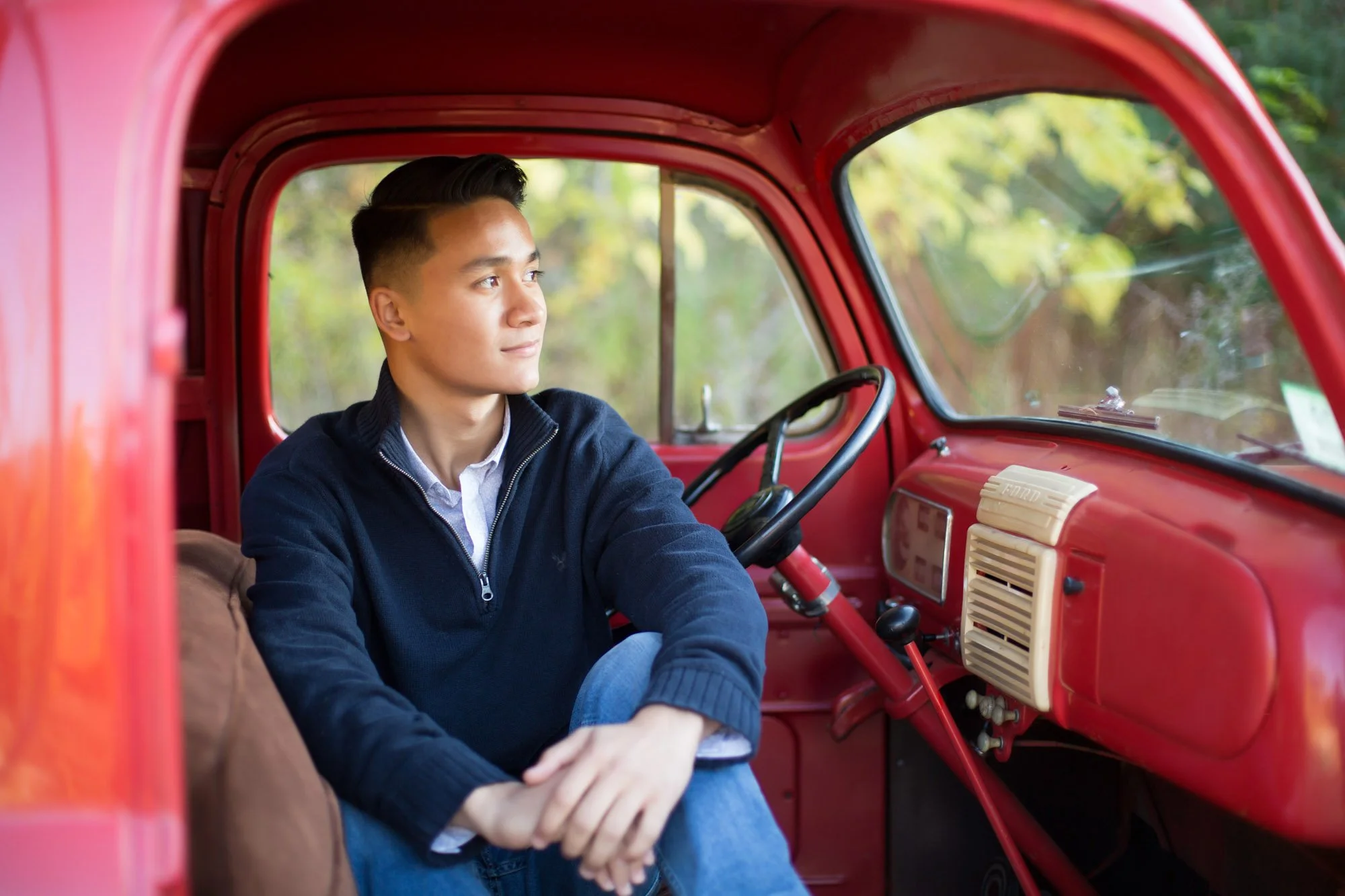 A young man sitting in the driver's seat of a red vintage truck, looking out the window with a peaceful expression, with green foliage visible outside.