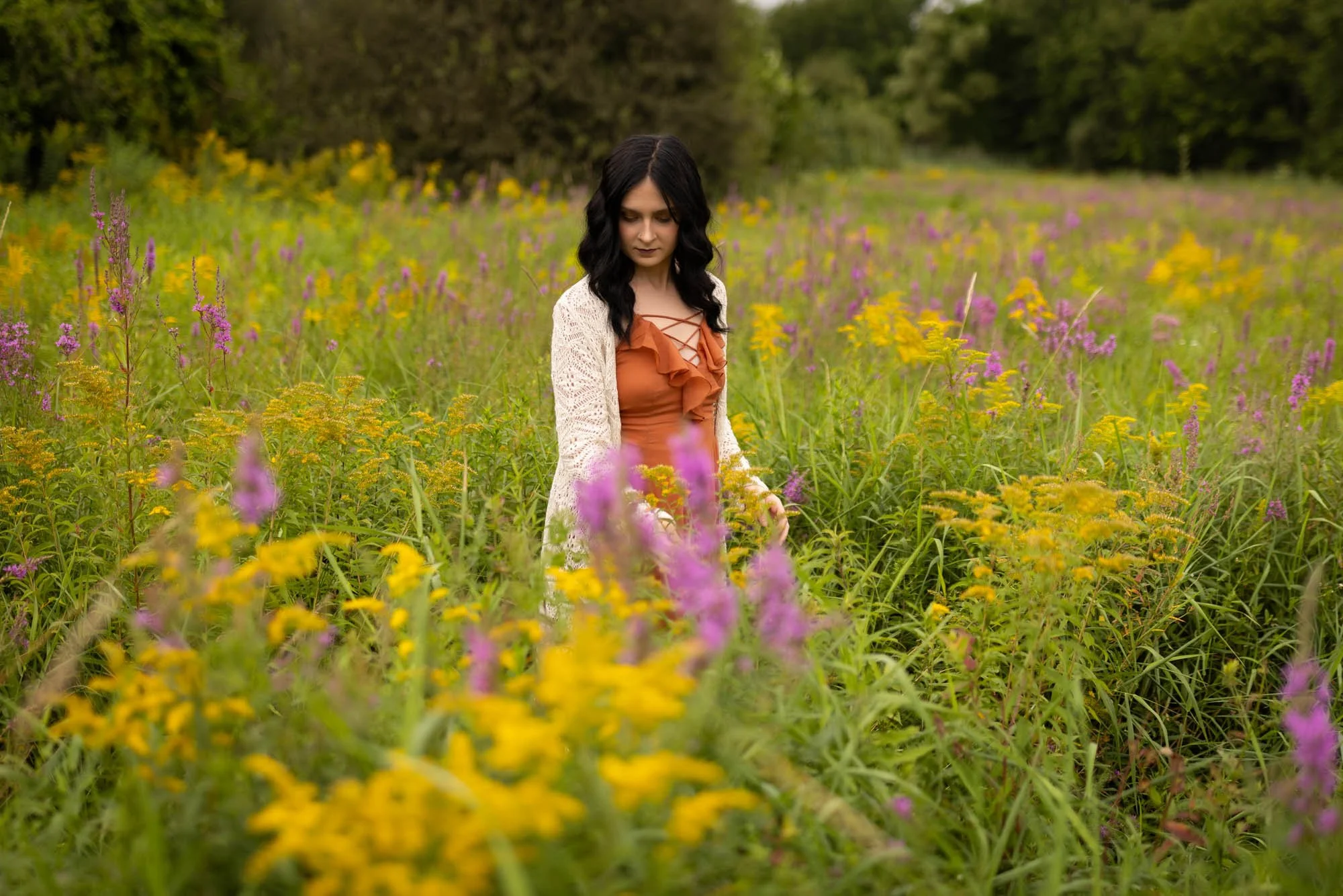 Woman with black hair in a rust-colored top and a white lace cardigan walking through a field of yellow and purple wildflowers.