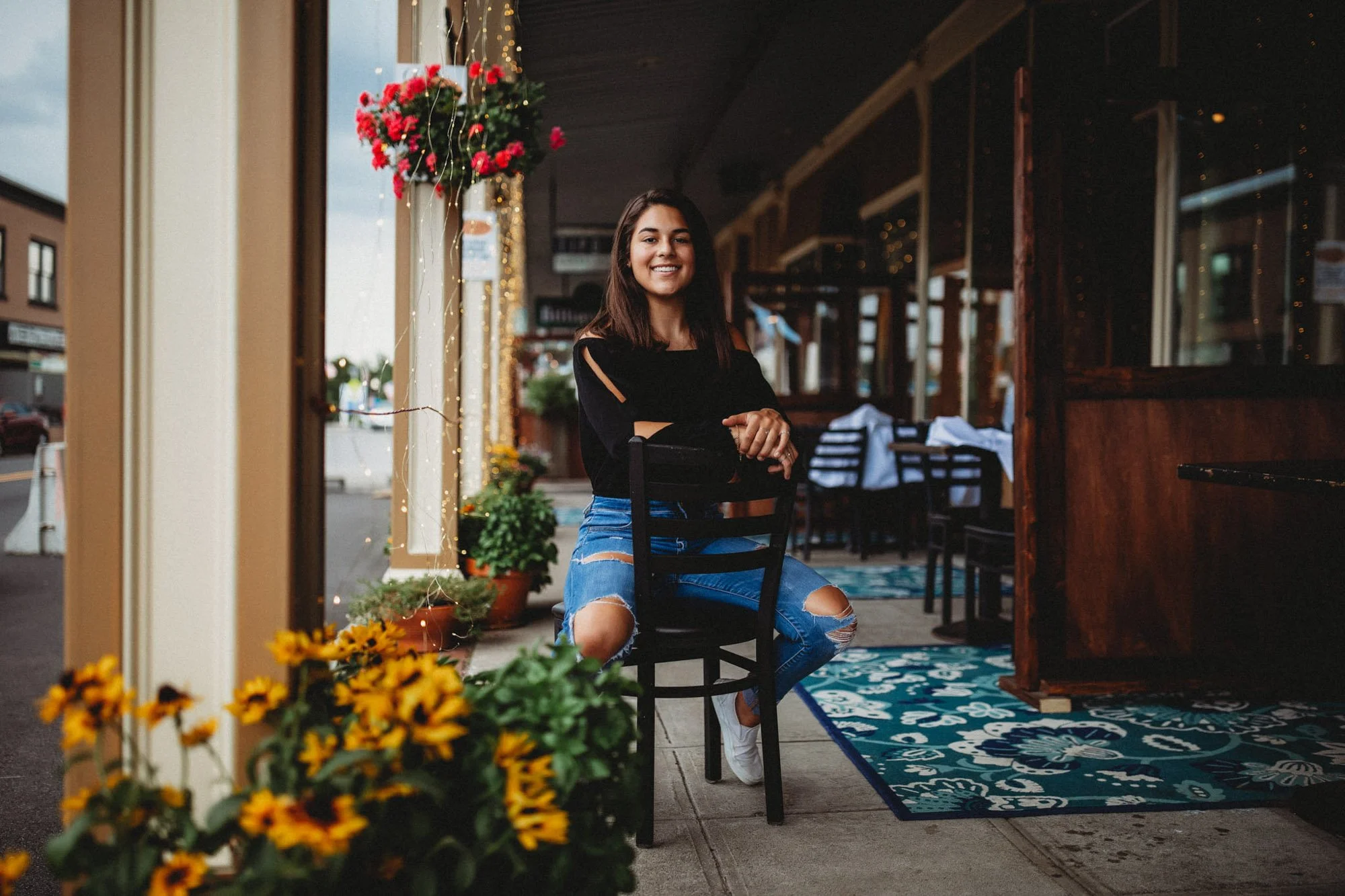 A young woman with dark hair, wearing a black top and ripped jeans, sitting on a black chair outside a restaurant with string lights, potted plants, and yellow flowers in the foreground