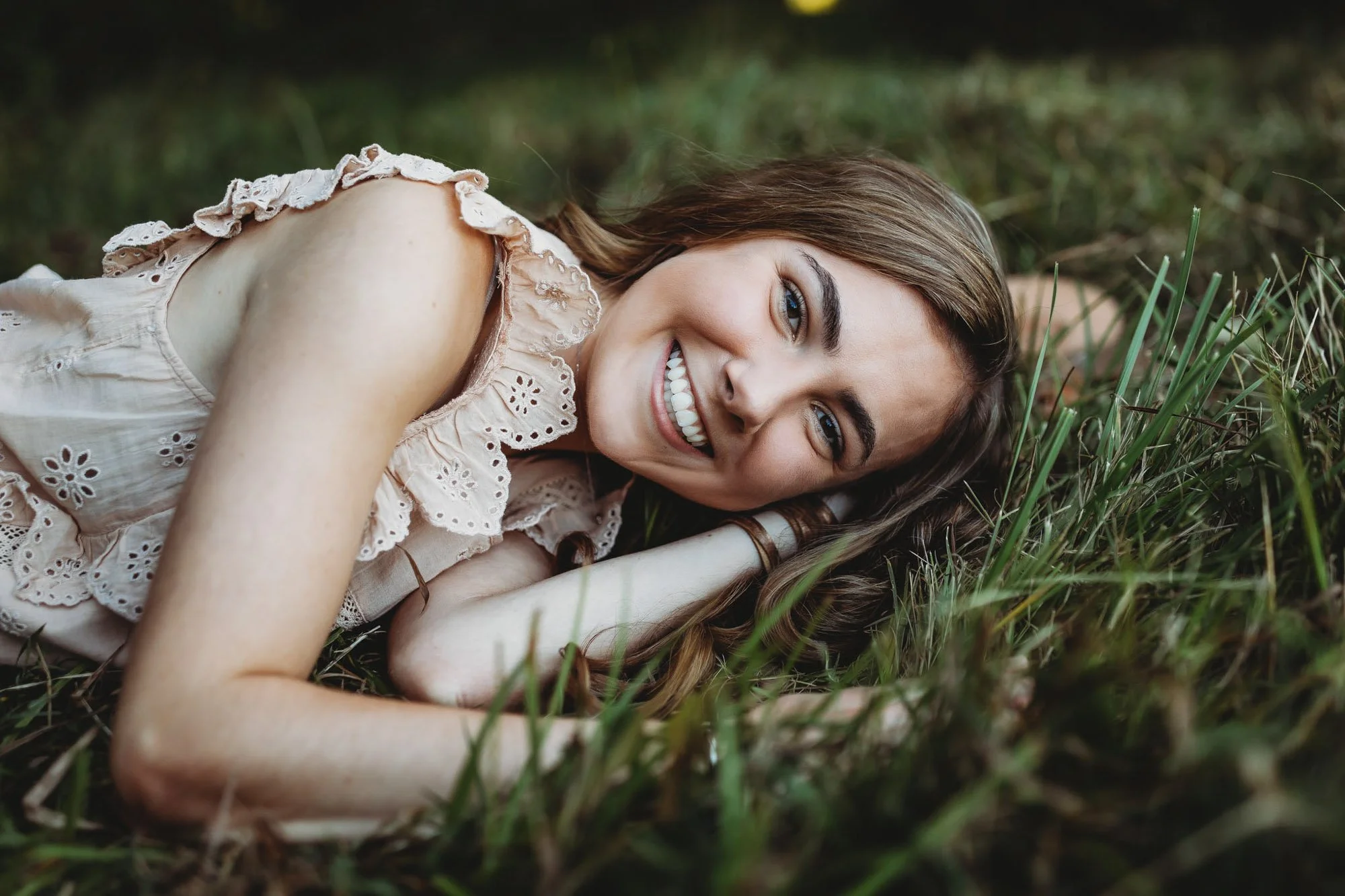A woman lying on the grass with a smile, wearing a light-colored eyelet blouse, and looking at the camera.