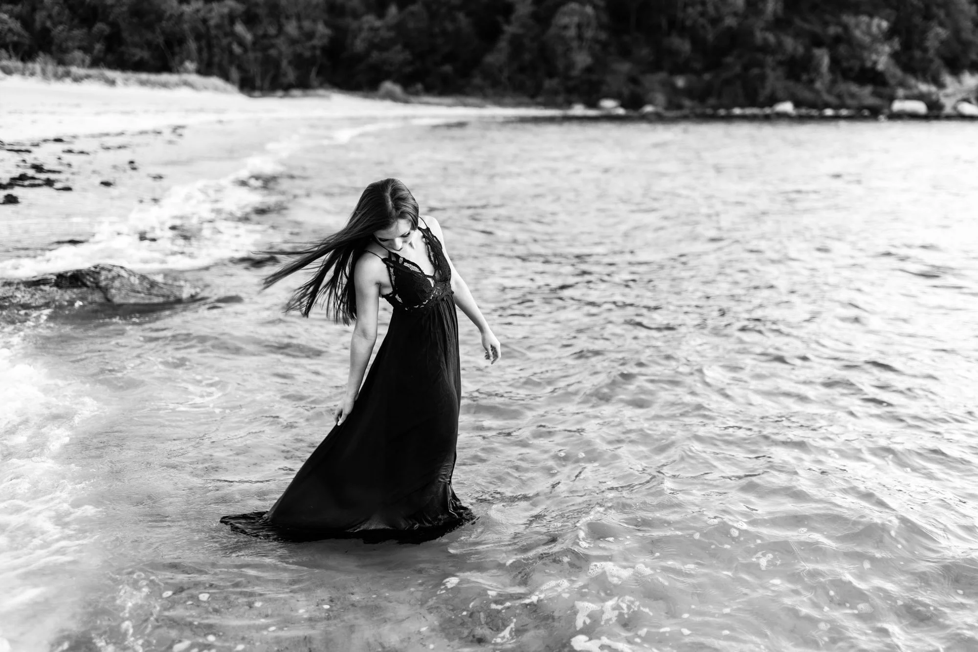 A woman in a long black dress standing by the shoreline, with her head bowed and hair flowing, near the water's edge.