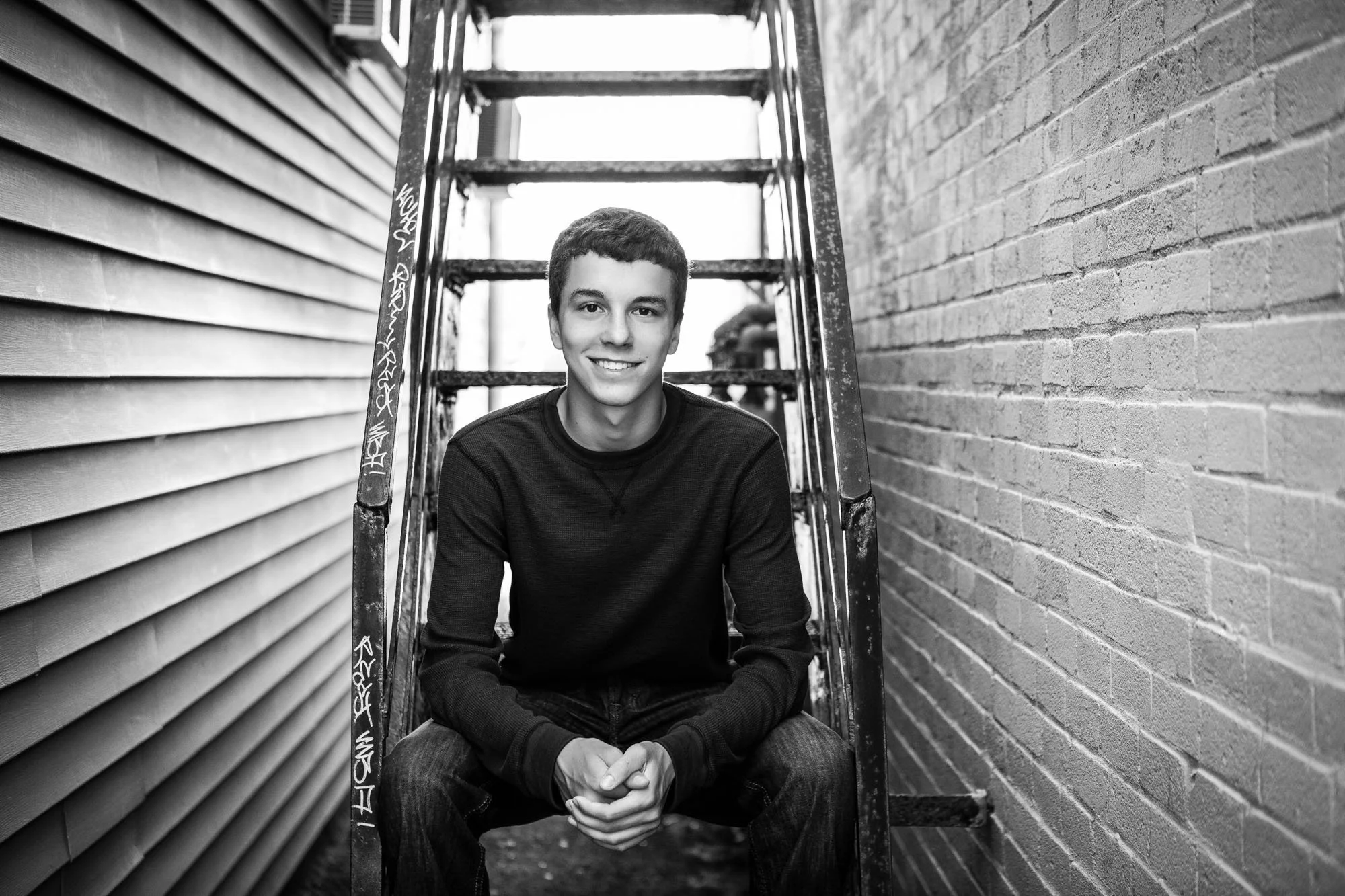 A young man with short dark hair smiling, sitting on a step between a brick wall and a building with horizontal siding, in an outdoor urban alleyway.