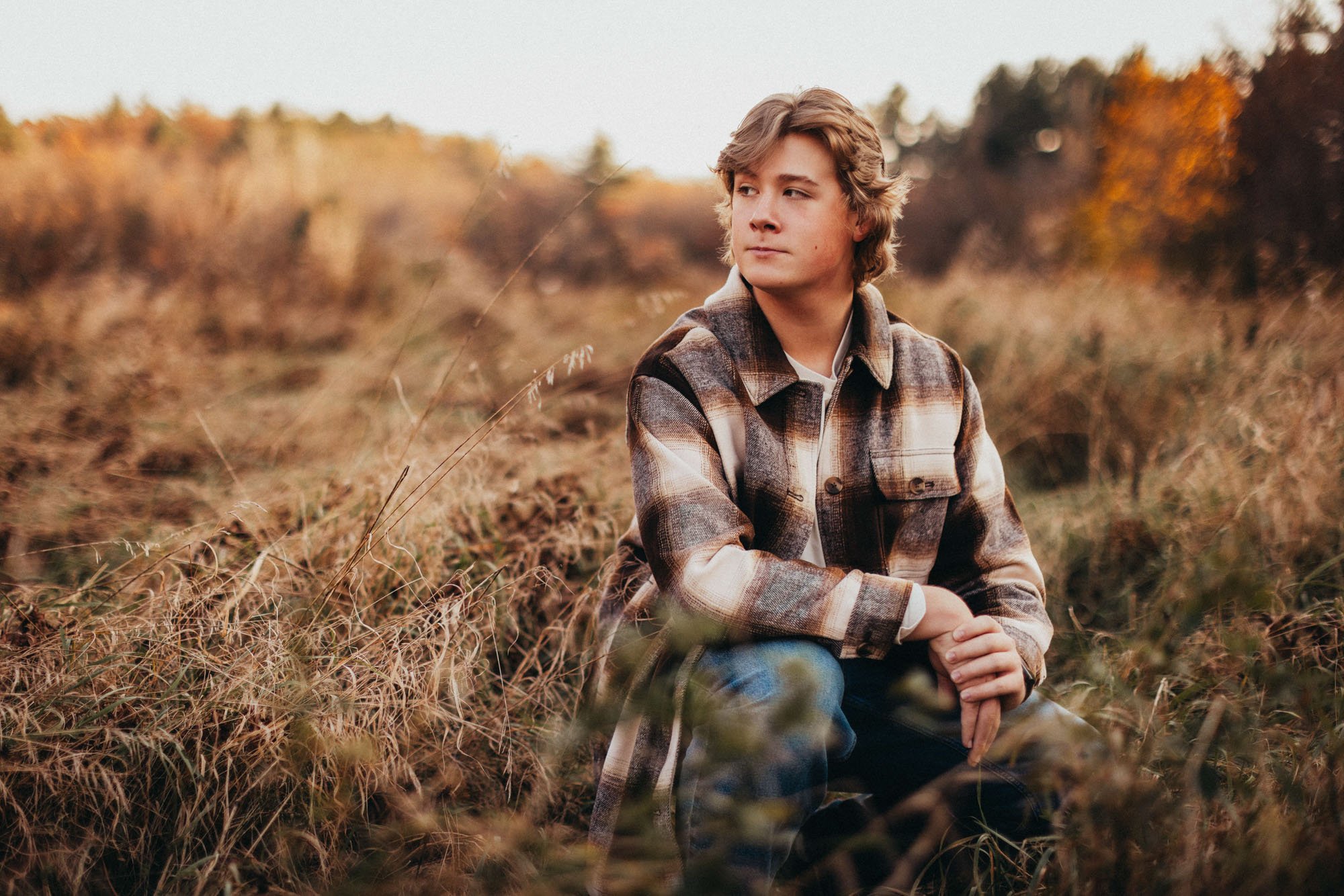 A young man in a plaid jacket sitting in a field of dry grass during autumn, with trees showing fall colors in the background.