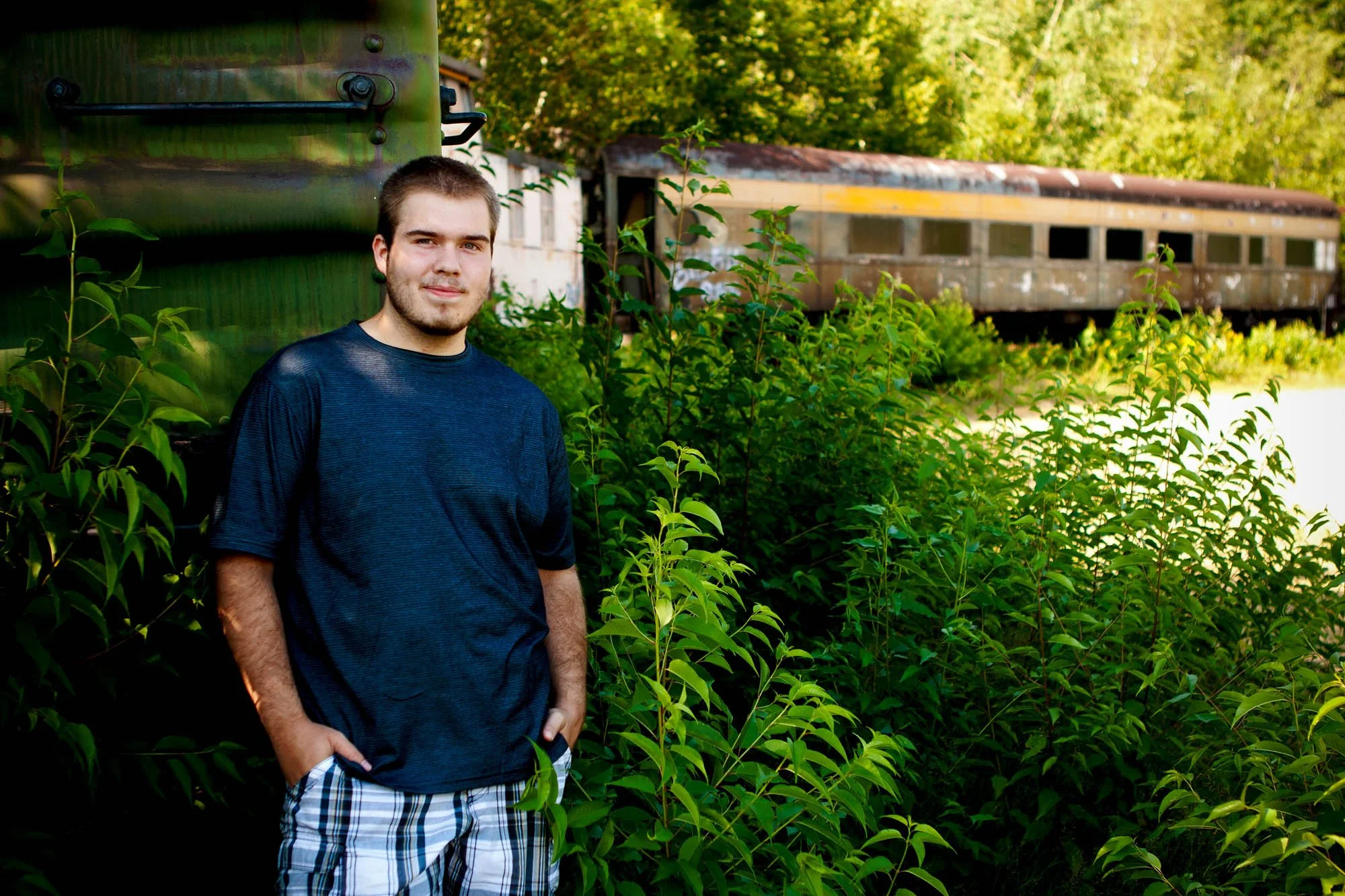 Young man standing outdoors with green plants and an old train car in the background.