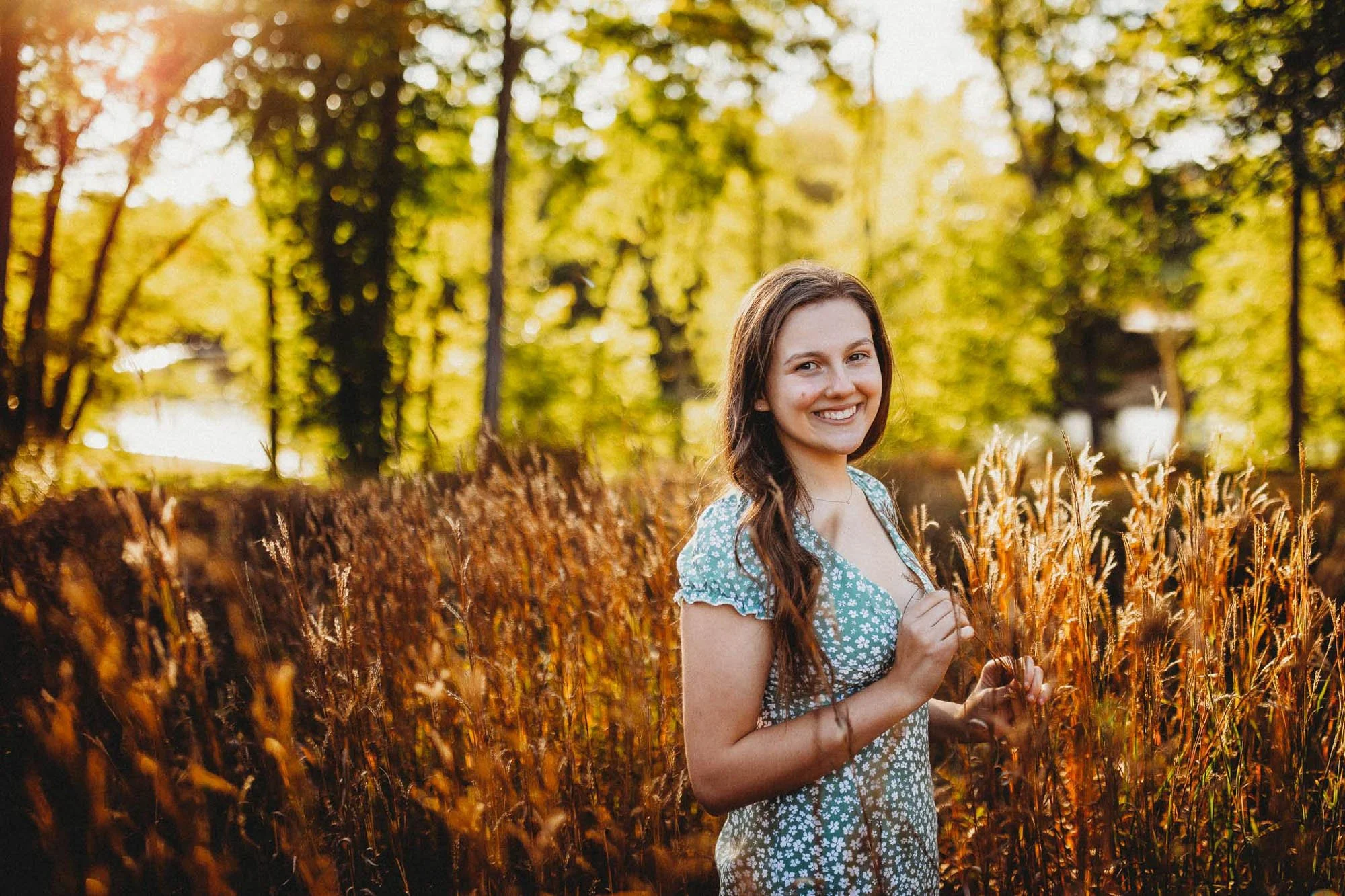 A young woman with long brown hair smiles while standing in a field of tall, brown grass during golden hour, with trees and sunlight in the background.
