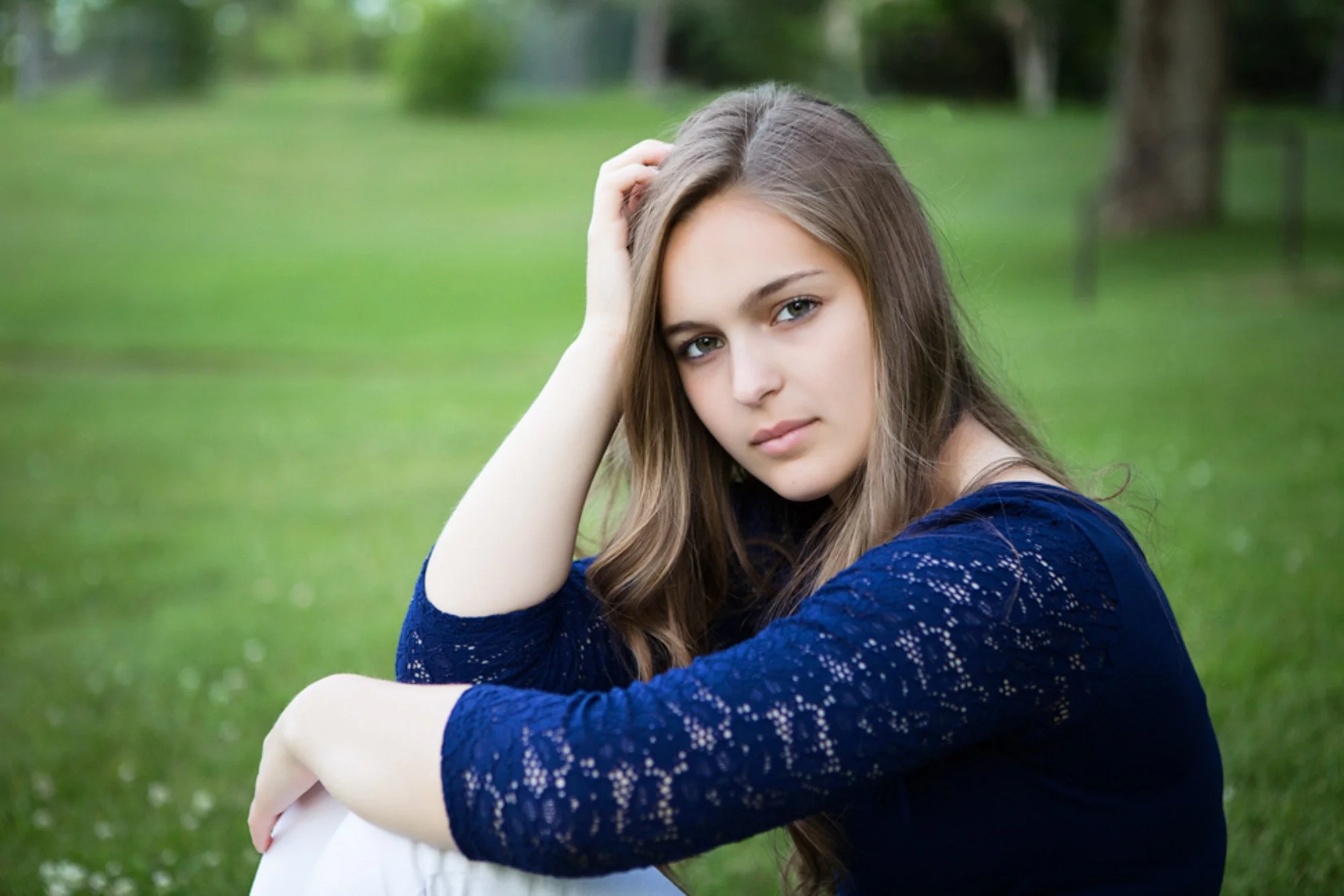 Young woman with long brown hair sitting outdoors on grass, wearing a navy blue lace top, looking into the camera with a contemplative expression, green trees in the background.