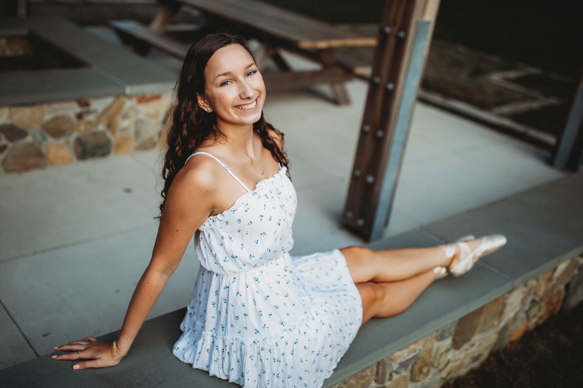 A young woman in a white sundress with small blue flowers sitting on a concrete ledge. She has curly brown hair and is smiling at the camera. The background includes a stone wall and a wooden structure.