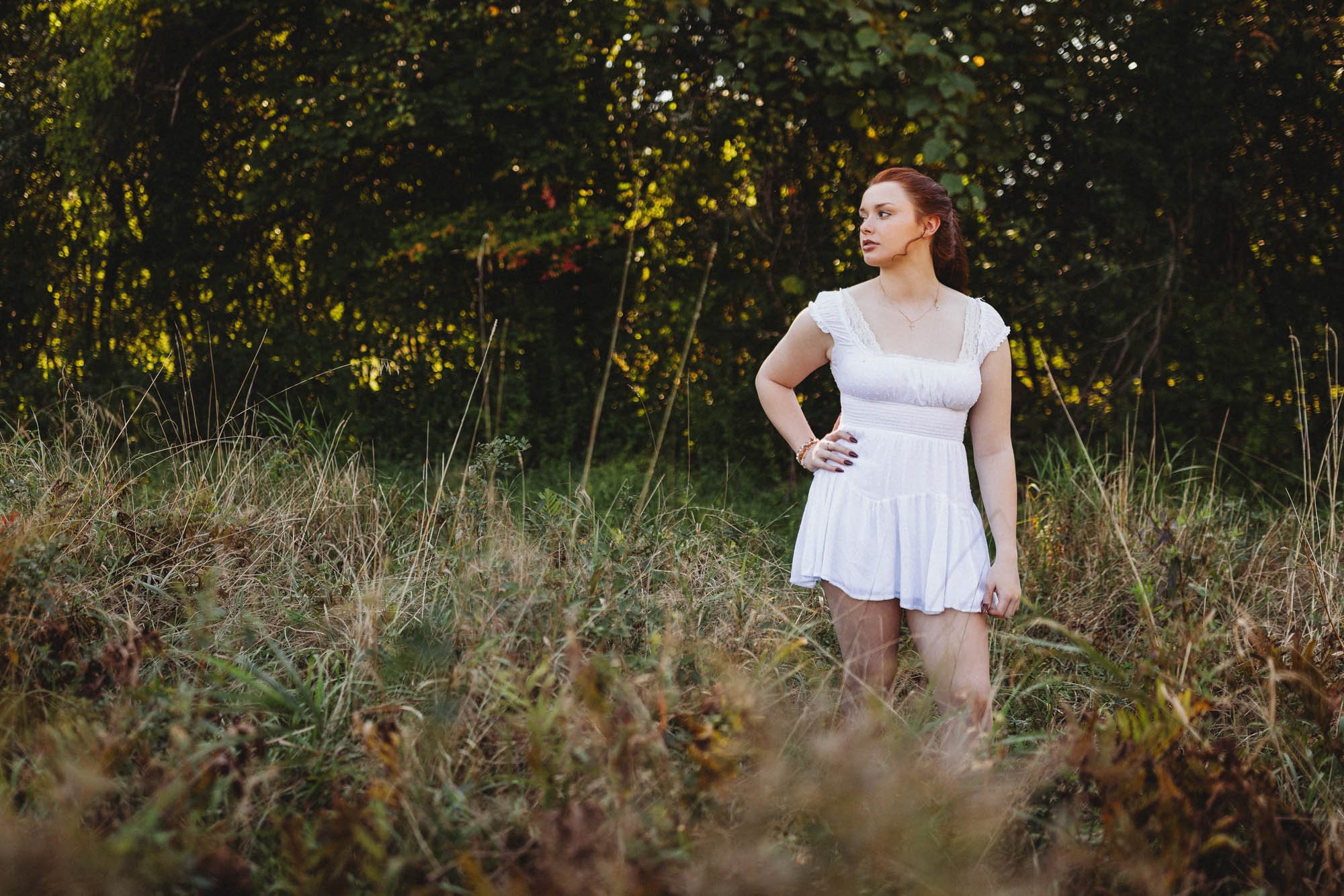 Woman in a white dress standing in a grassy field with trees in the background.