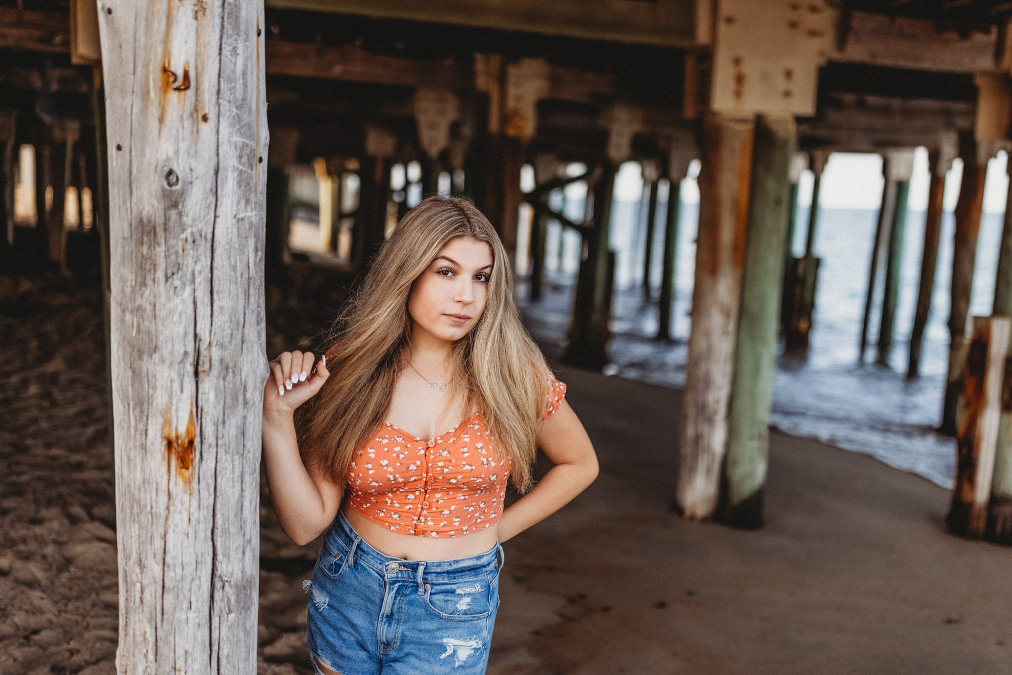 A young woman with long blonde hair standing under a pier at the beach, wearing a reddish-orange crop top with white floral pattern and denim shorts.