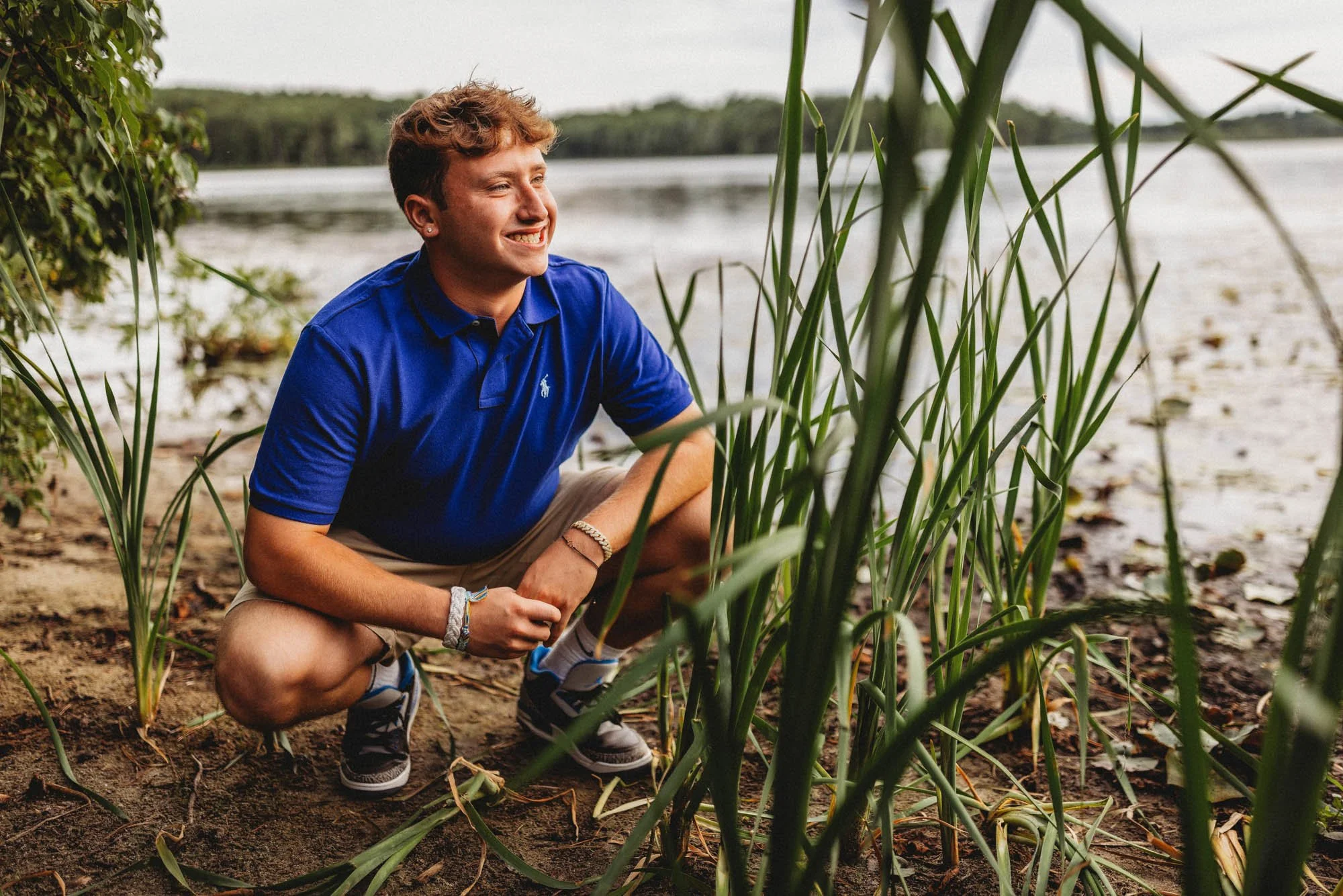 A young man crouching near the edge of a lake, surrounded by tall grass and trees in the background, smiling and looking attentively to his right.