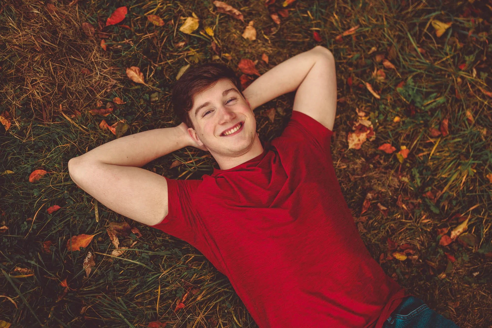 Young man lying on the grass with autumn leaves, smiling and with arms behind his head, wearing a red t-shirt.