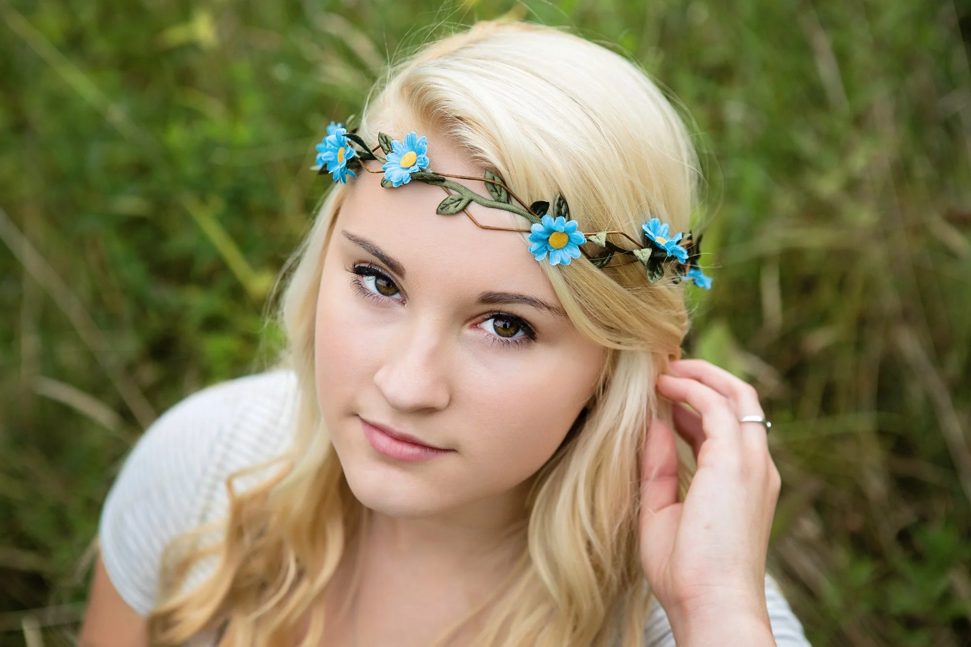 A young woman with long blonde hair wearing a blue flower crown, outdoors with green foliage in the background.