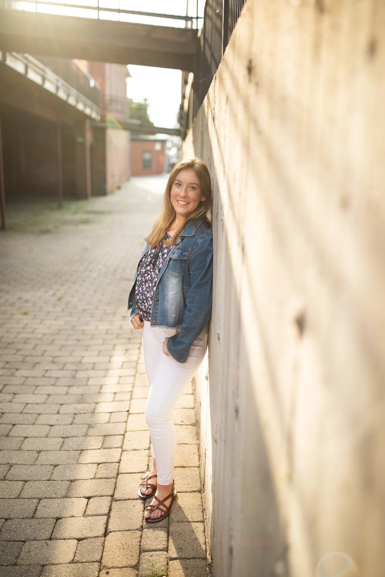 Young woman with long light brown hair leaning against a wooden wall in an outdoor alleyway, smiling at the camera, wearing a denim jacket, floral top, white pants, and sandals, with sunlight creating a warm glow.