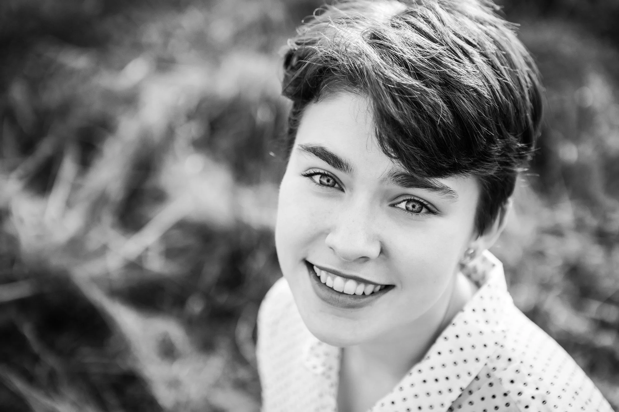 Black and white portrait of a young woman with short, dark hair and light-colored eyes, smiling outdoors.