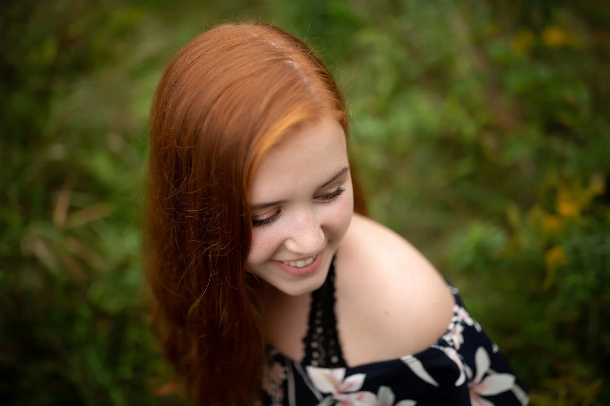 A young woman with long red hair smiling and looking down in an outdoor setting with green foliage.