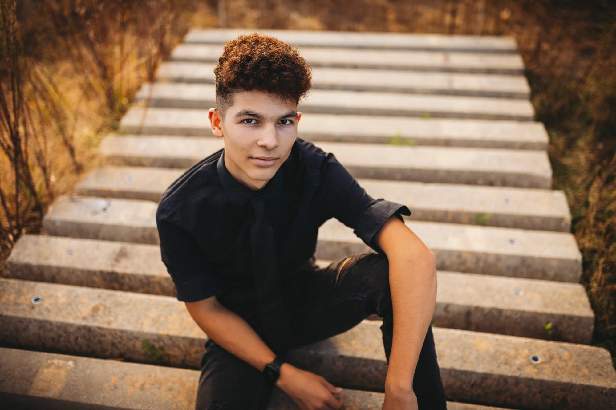 Young man with curly hair, wearing a black shirt and black pants, sitting on outdoor stone steps with grass on the sides.