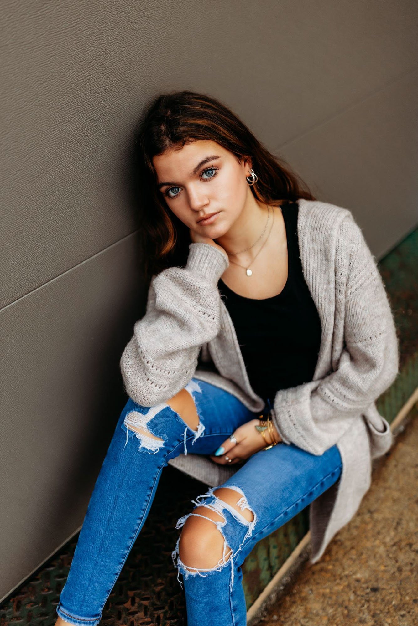 Young woman with dark hair, blue eyes, and multiple earrings sitting against a wall, wearing a black top, a light gray cardigan, and ripped blue jeans.