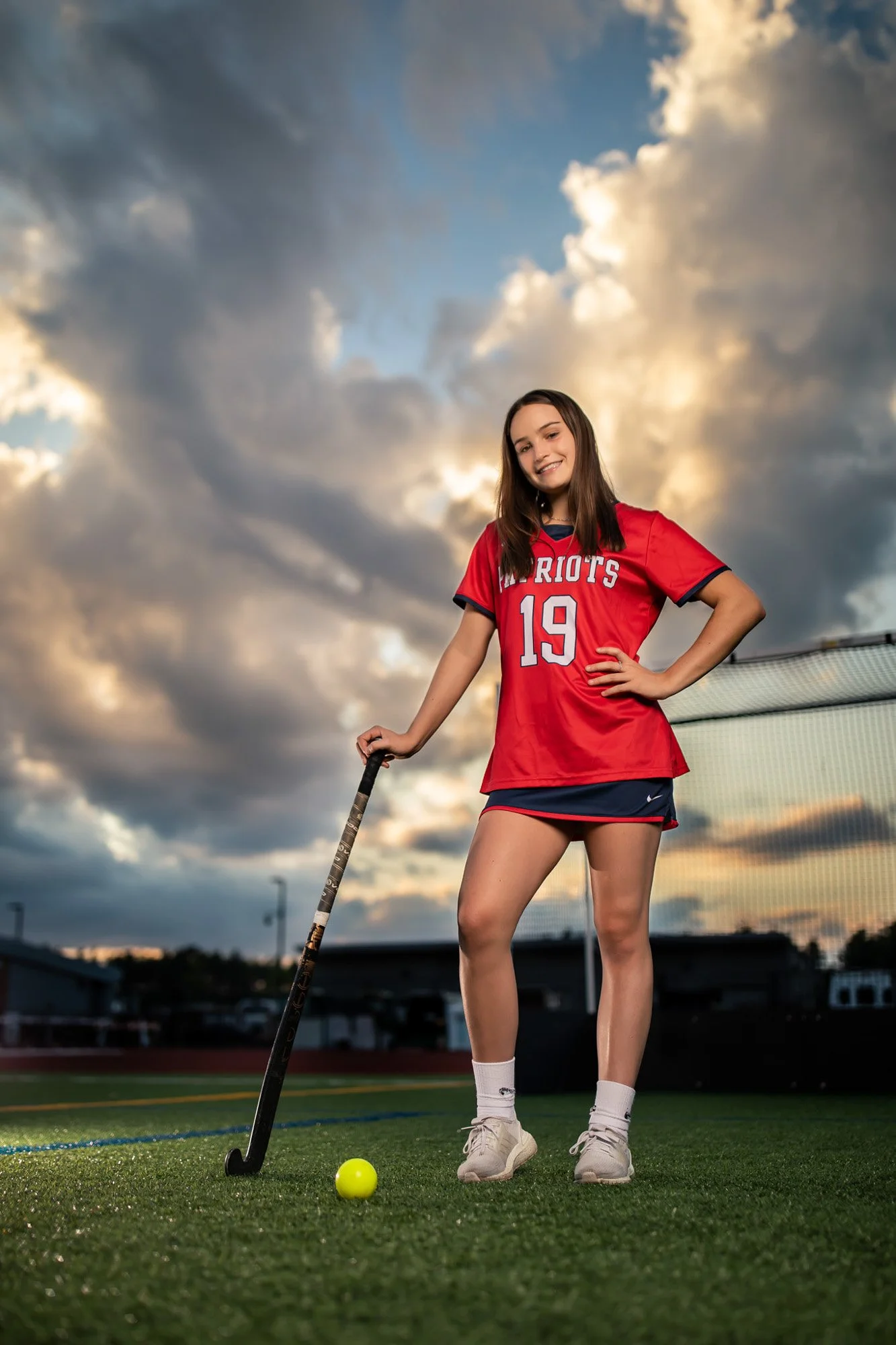 Young girl in a red sports uniform standing on a field at sunset, holding a hockey stick and smiling, with a tennis ball nearby.