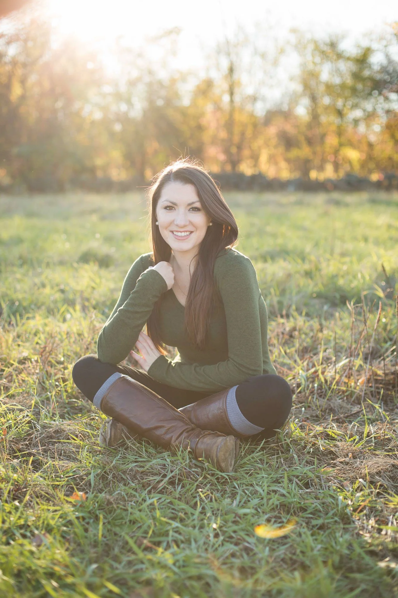 Young woman with long brown hair sitting cross-legged in a grassy field during golden hour, smiling at the camera with autumn trees in the background.