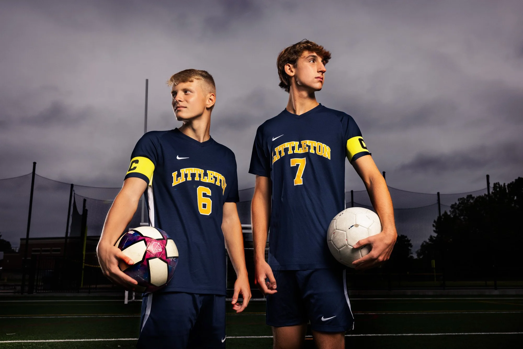 Two young male soccer players in dark blue uniforms with yellow lettering that says Littleton, each holding a soccer ball, standing on a field with a stormy sky in the background.