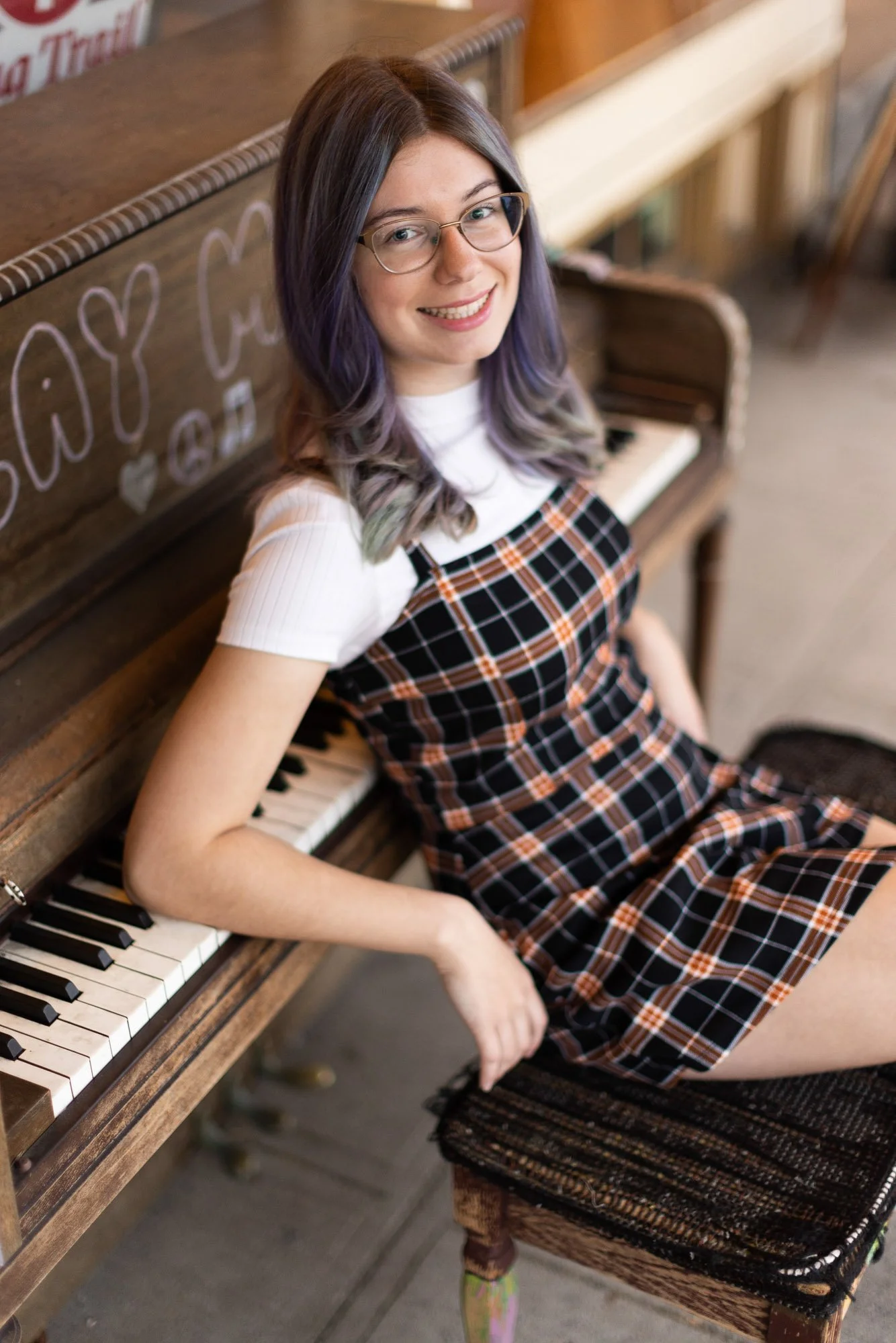 Young woman with glasses and multi-colored hair sitting on a stool next to a wooden piano, smiling at the camera.