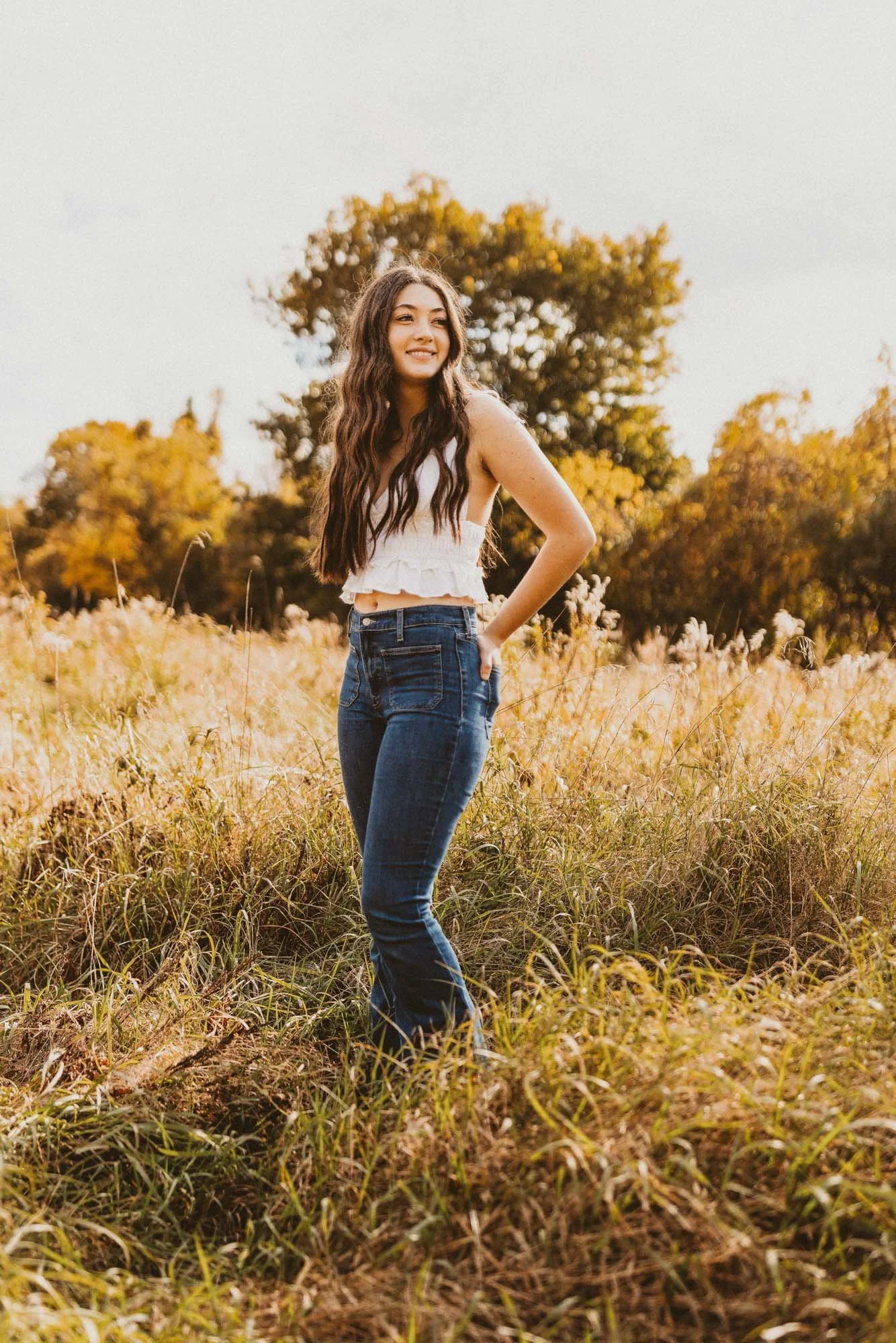Young woman with long wavy hair in a field during fall, smiling and wearing a white sleeveless top and blue jeans.