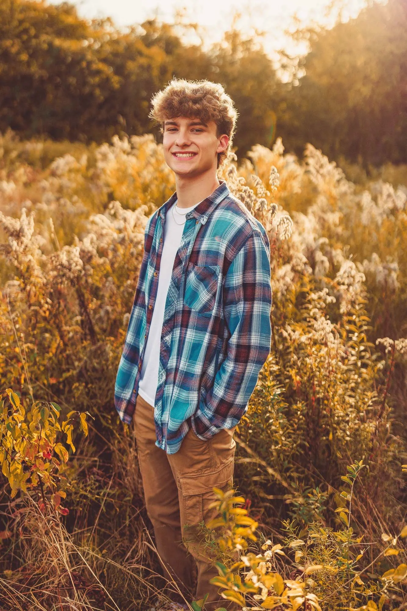 Young man smiling outdoors in a field of tall, golden plants during sunset, wearing a plaid shirt and beige pants.