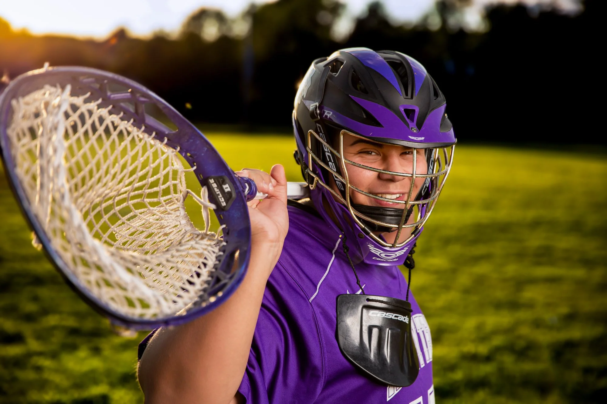 Lacrosse player wearing a purple helmet and jersey, holding a lacrosse stick over her shoulder in a grassy field at sunset.