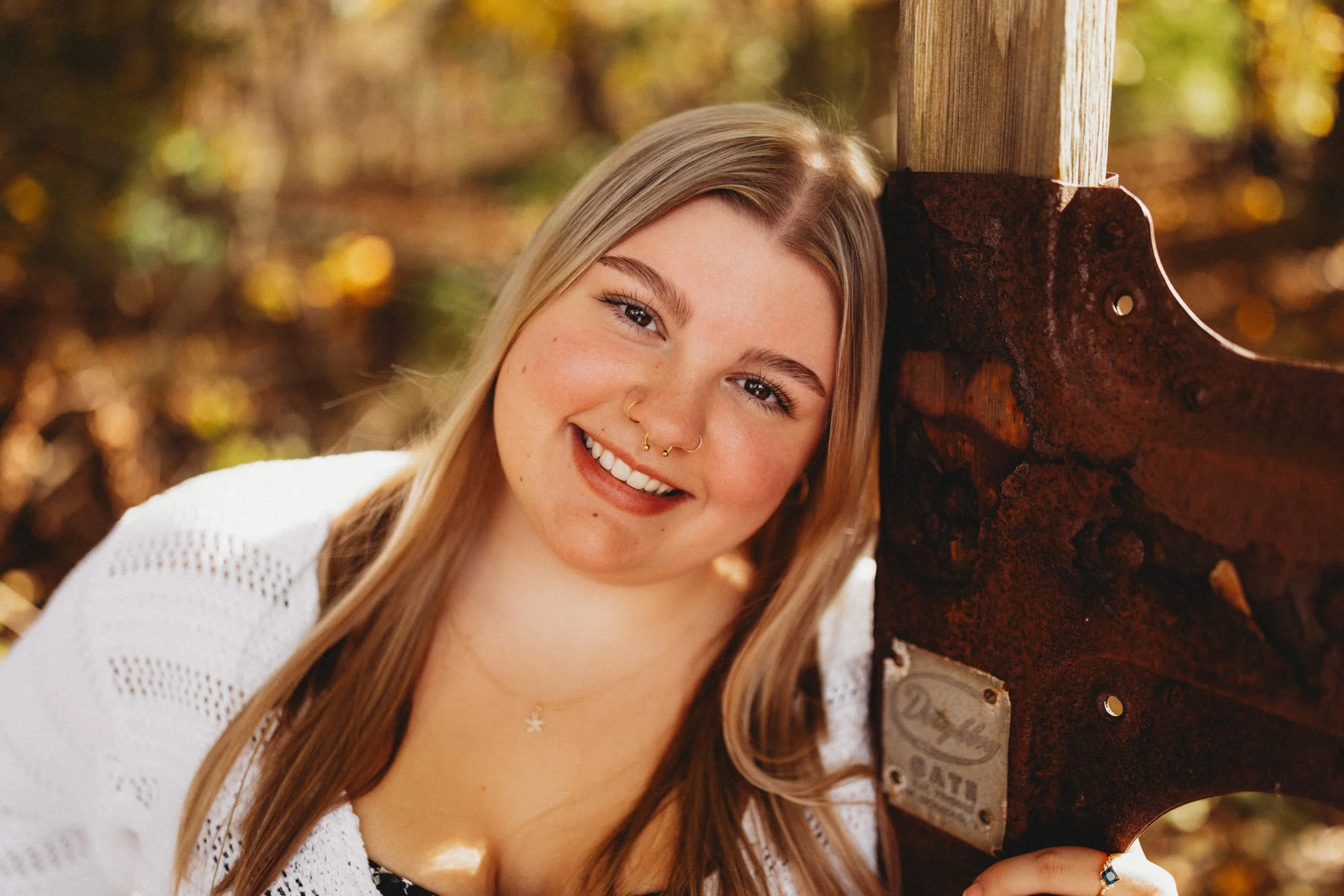 A young woman with long blonde hair, light skin, and multiple facial piercings smiling and leaning with her cheek against a rusty metal post in an outdoor setting with fall foliage.