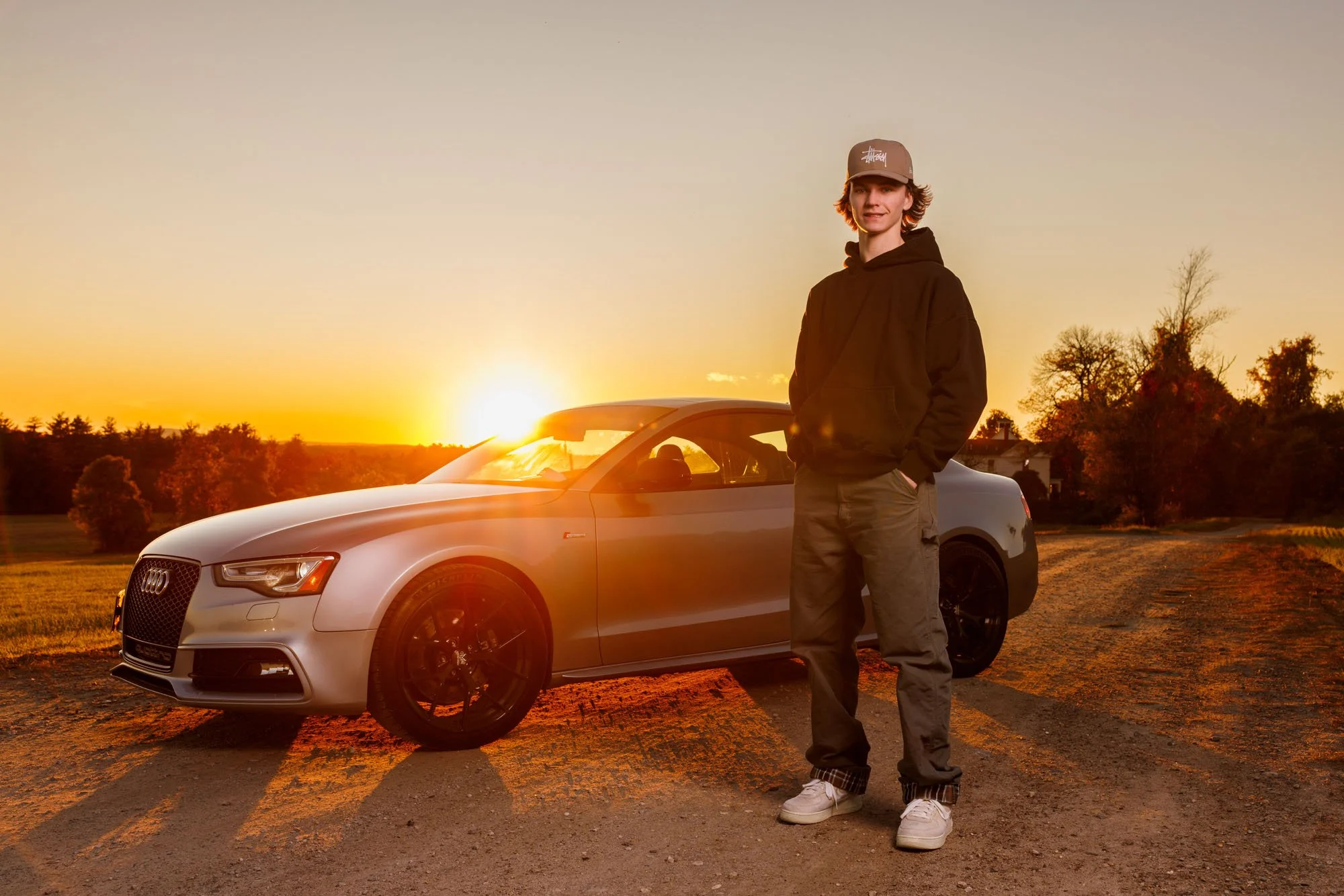 A young man standing beside a silver Audi car on a dirt road at sunset, wearing a black hoodie, gray pants, white sneakers, and a beige baseball cap.